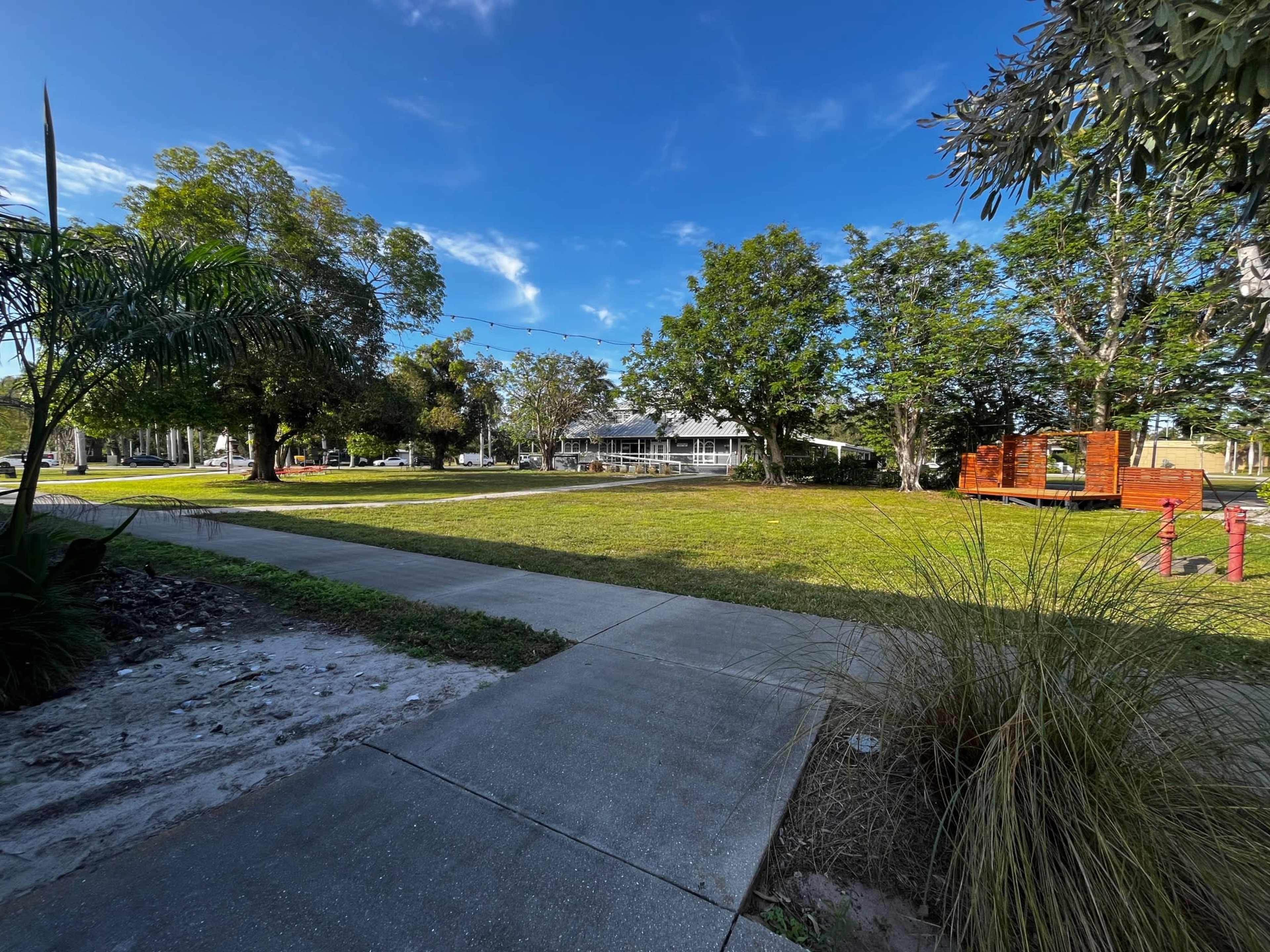 The image shows a grassy area with trees and a path, leading to a wooden structure in a park-like setting.