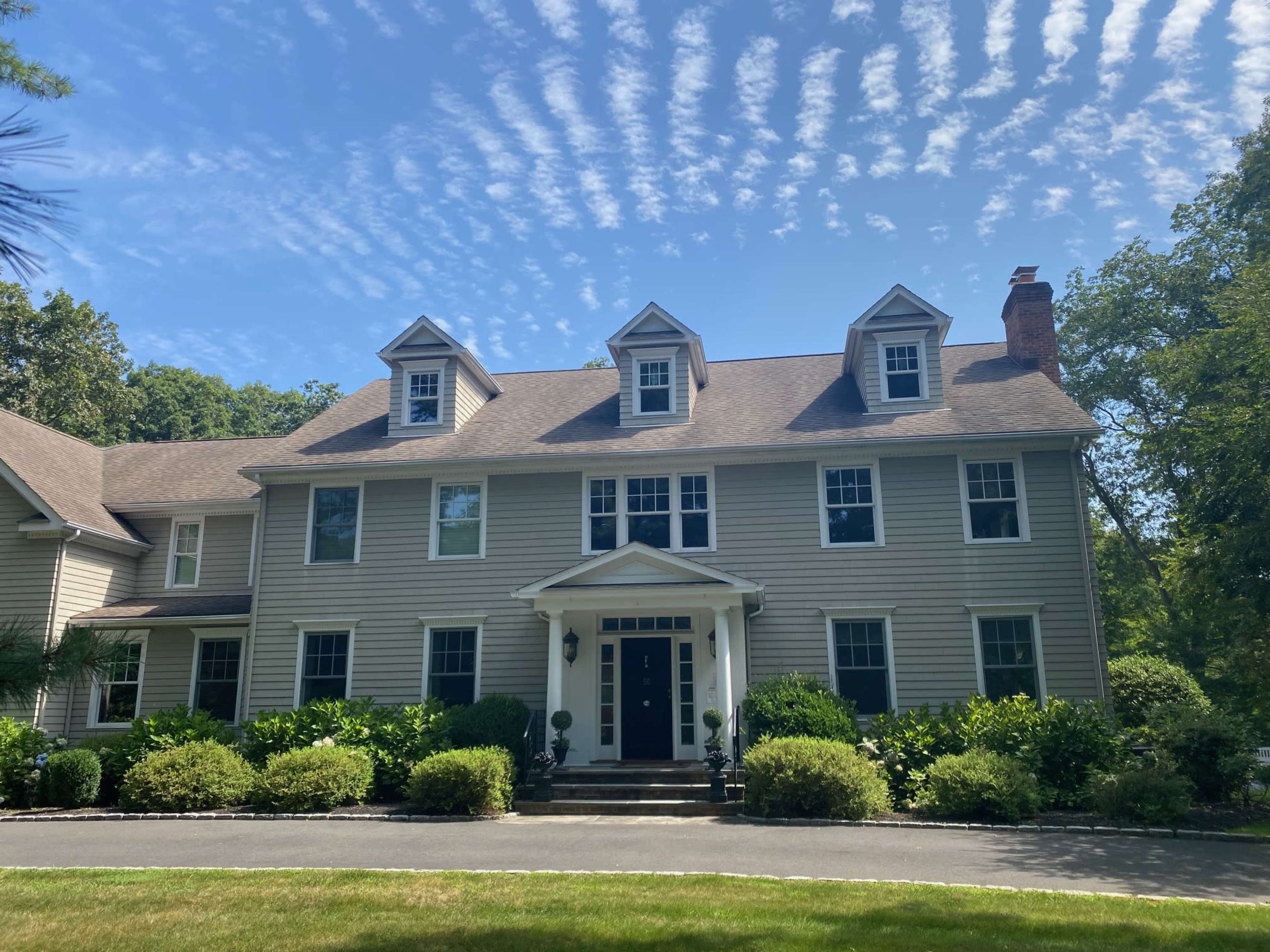 A large, two-story house featuring multiple gable roofs and a symmetrical facade is set against a backdrop of blue skies and wispy clouds.