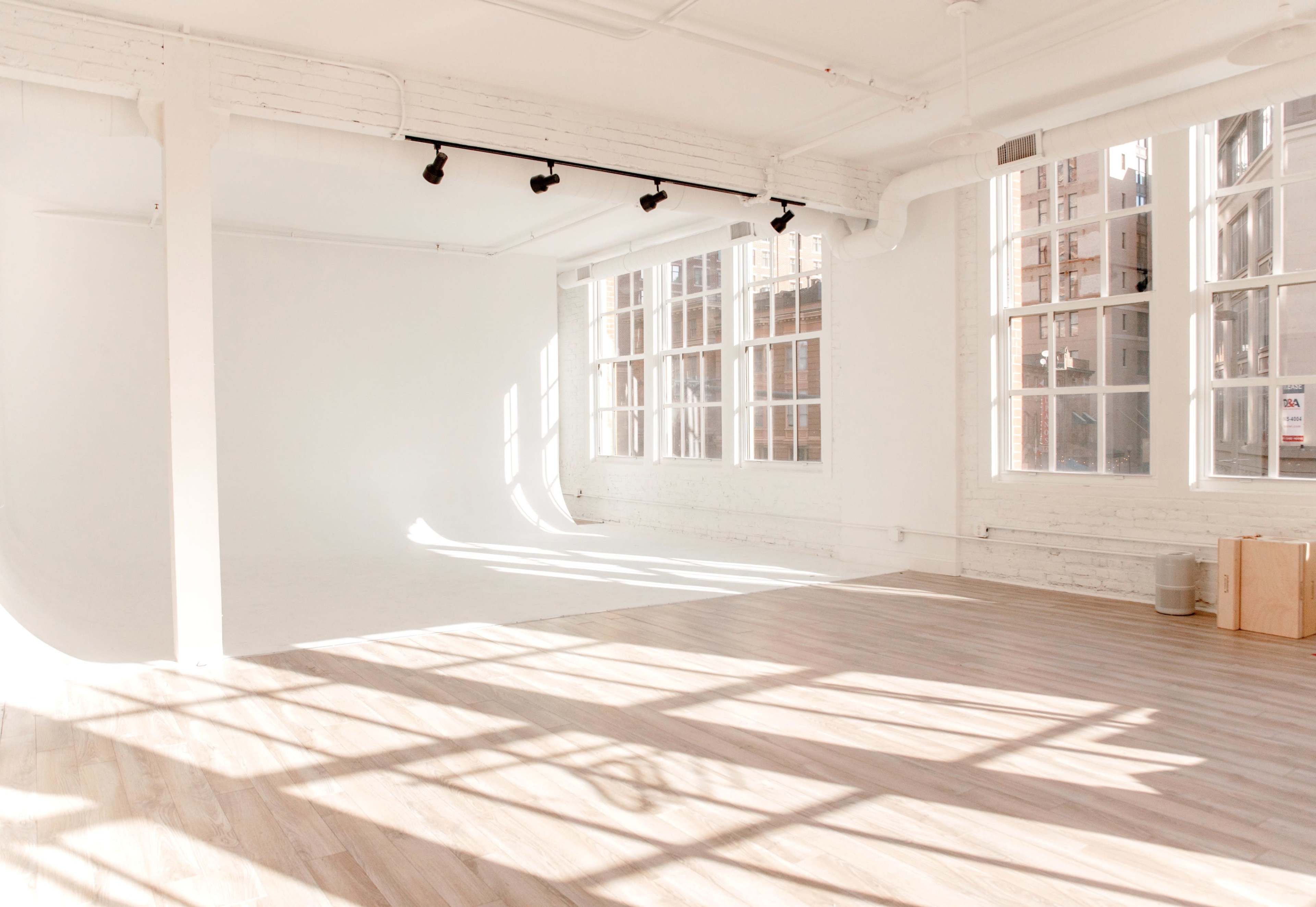 A spacious, empty room with large windows, white walls, and wooden flooring, illuminated by natural light.