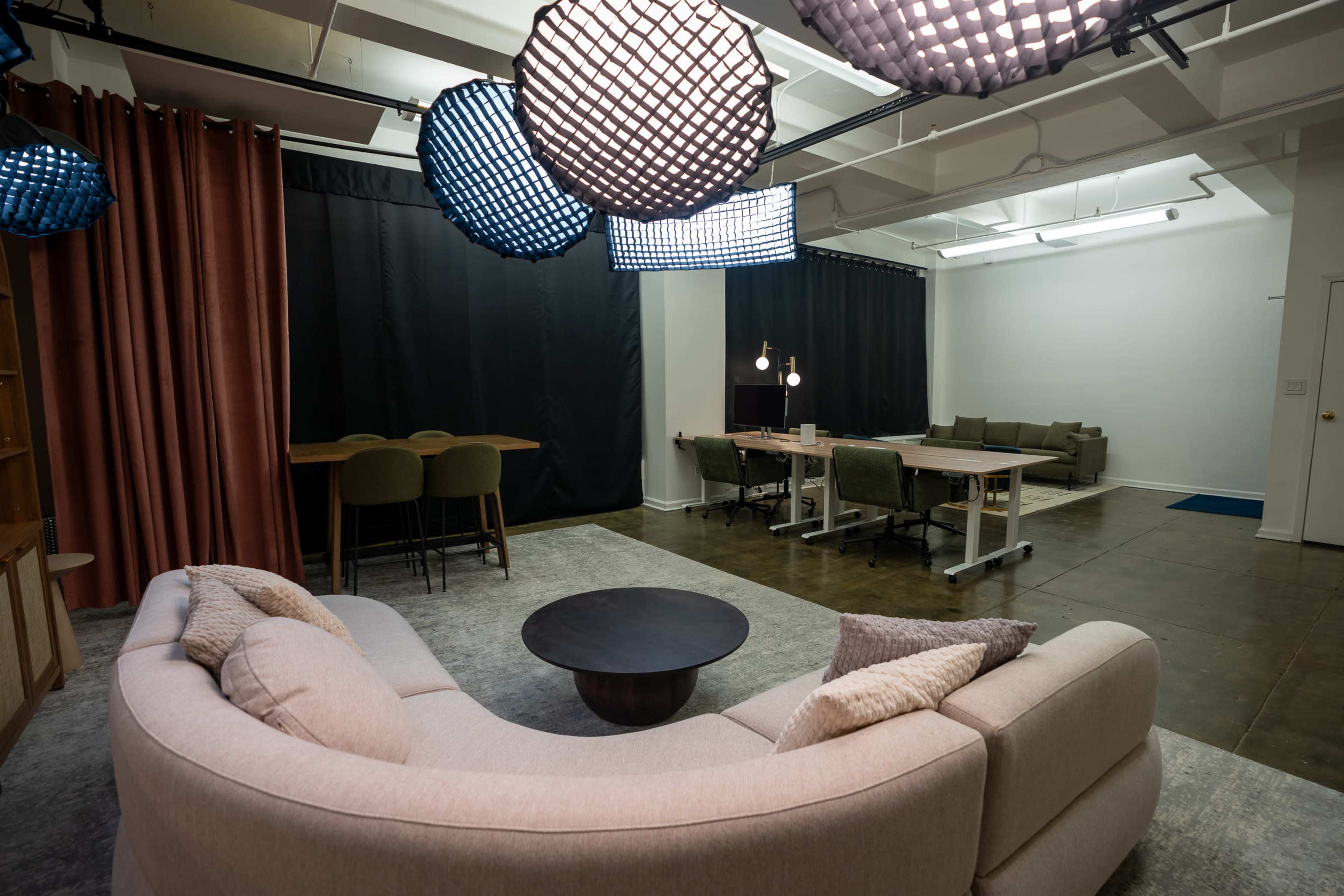 The image shows a modern lounge area with a curved sofa, a round coffee table, and several seating options in a well-lit, spacious room divided by black curtains.