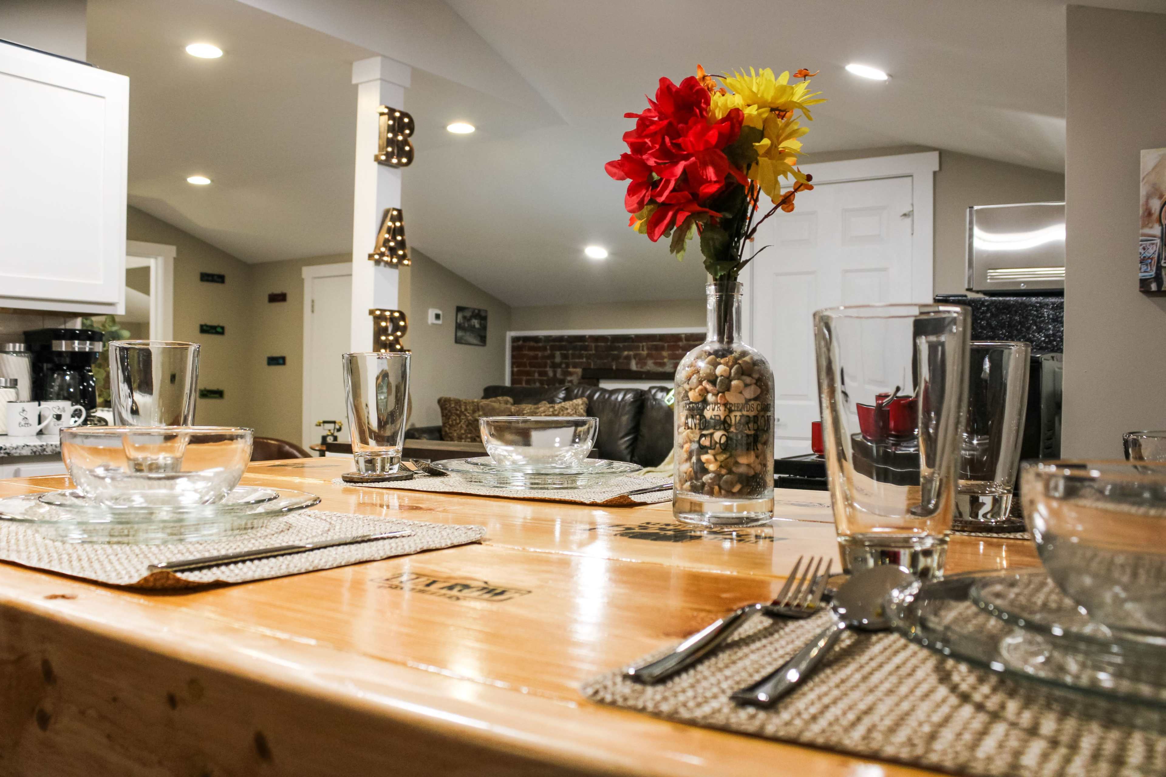 A dining table is set with glassware and utensils, featuring a vase of flowers and a rustic wooden surface, against a backdrop of a cozy living area.