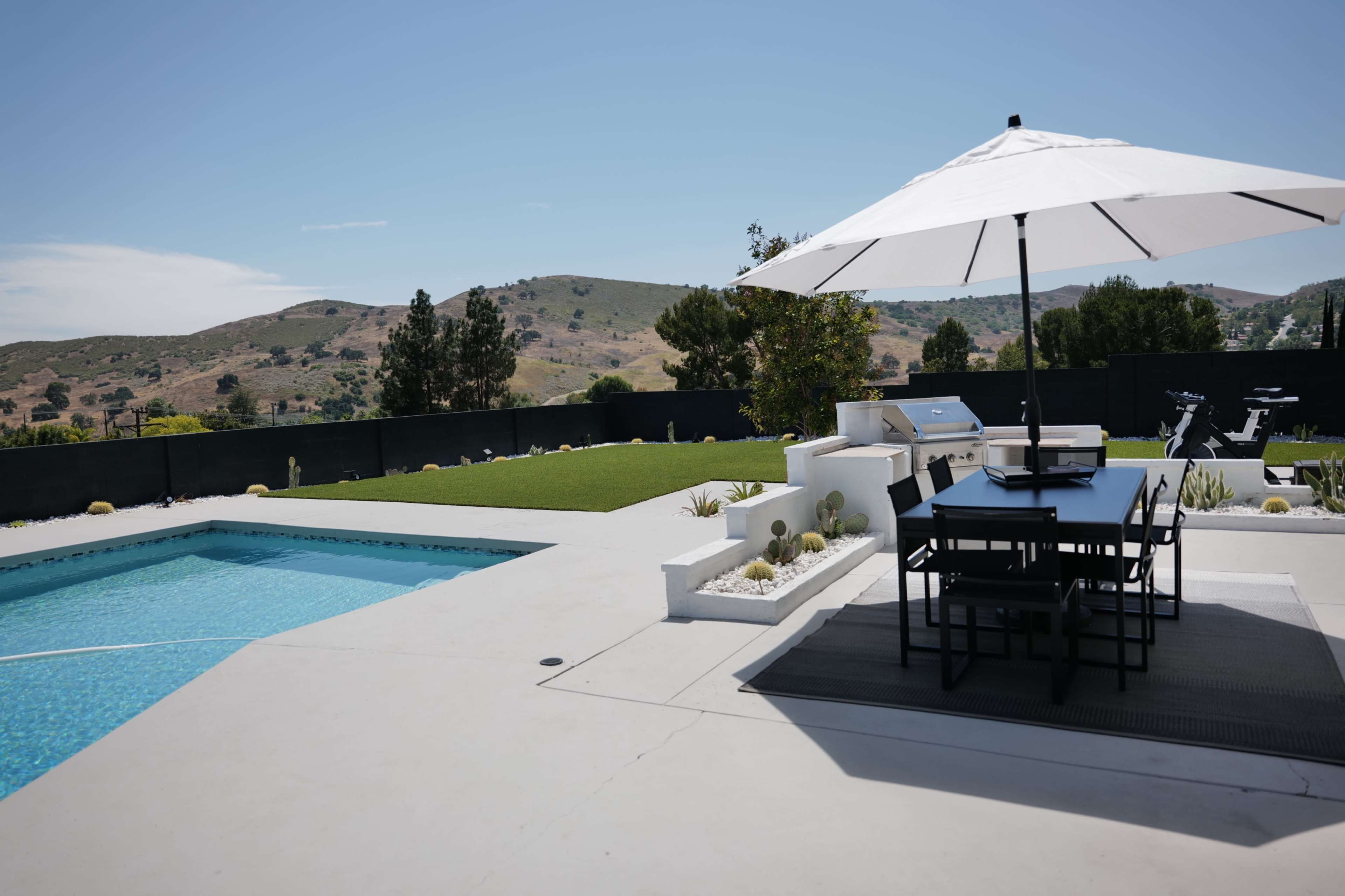 A large outdoor patio features a swimming pool, a dining table with chairs, and an umbrella, overlooking a green landscape and hills in the background.