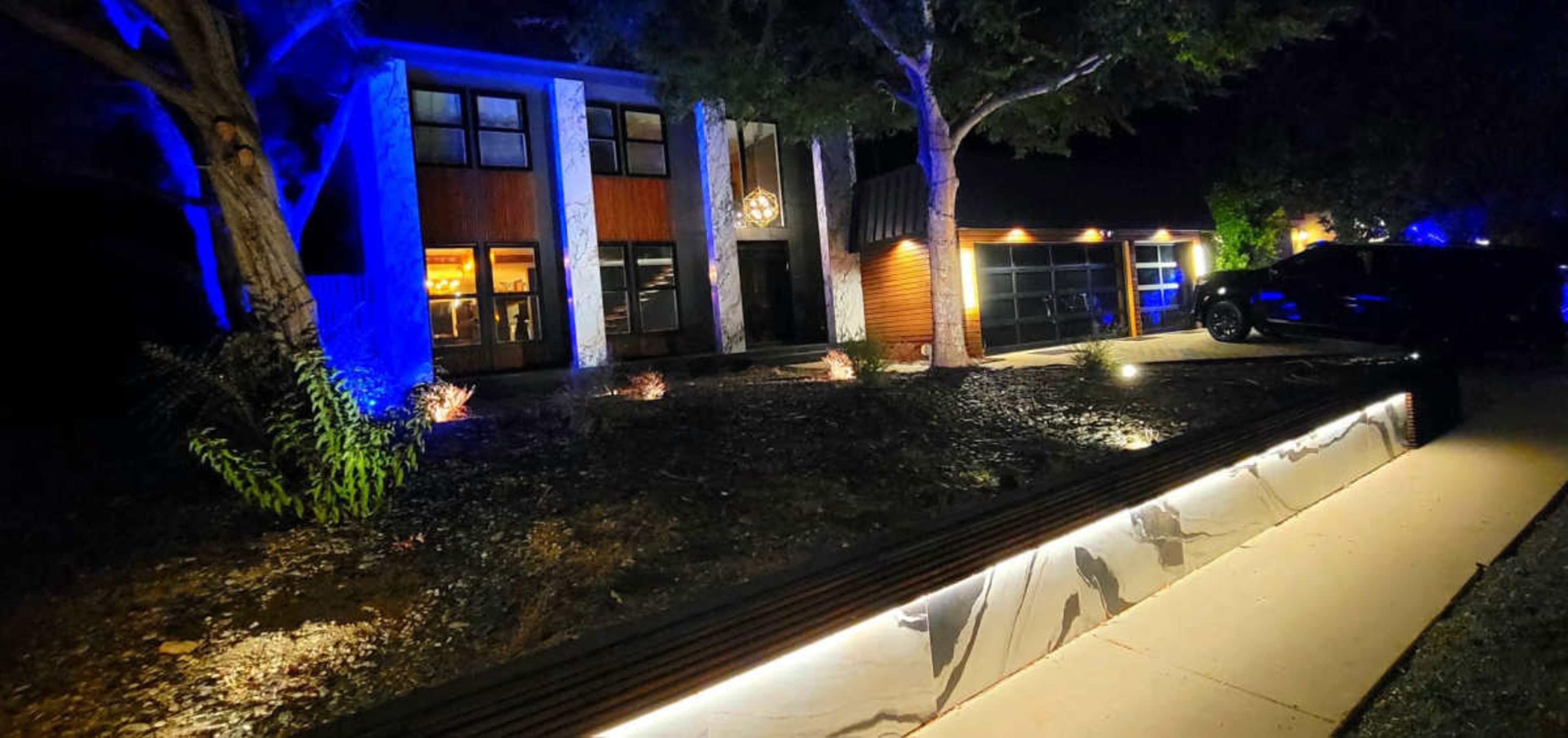 A modern two-story house is illuminated at night, featuring large windows and a well-lit driveway with landscaping.