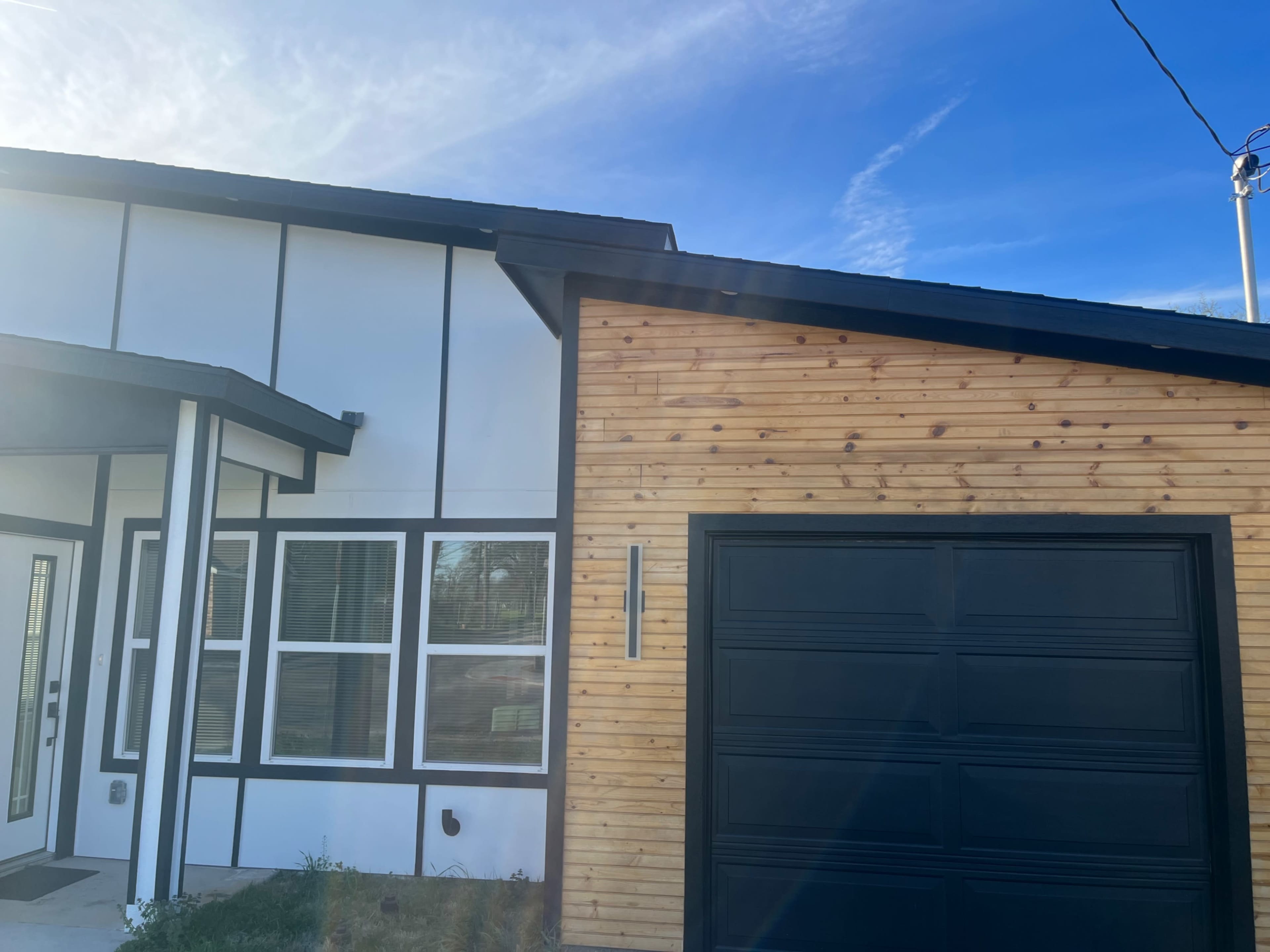 The image shows a modern house with a combination of white and wooden siding, featuring large windows and a black garage door.