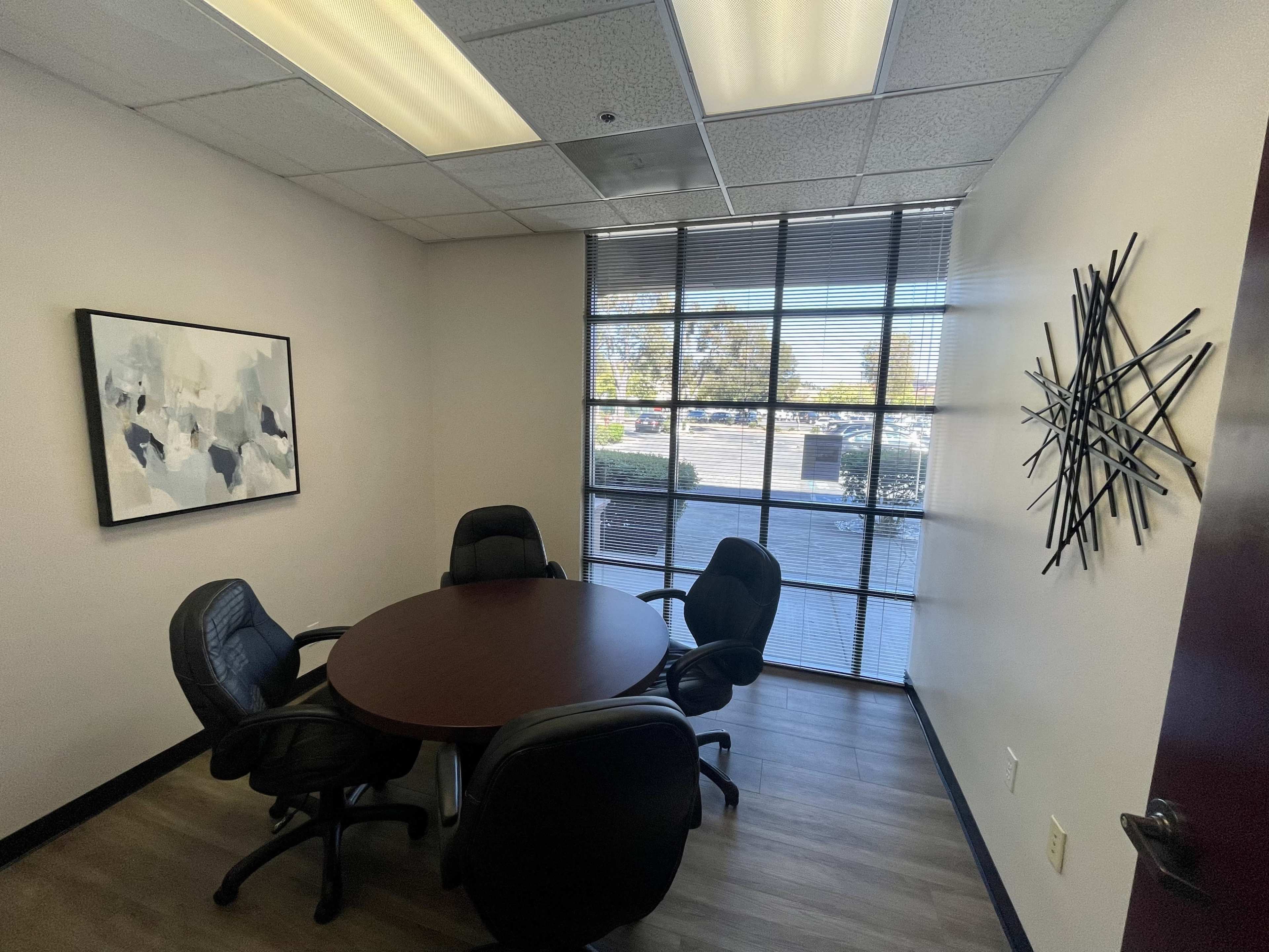 A small conference room features a round table surrounded by four black chairs and a large window with a view of an outside area.