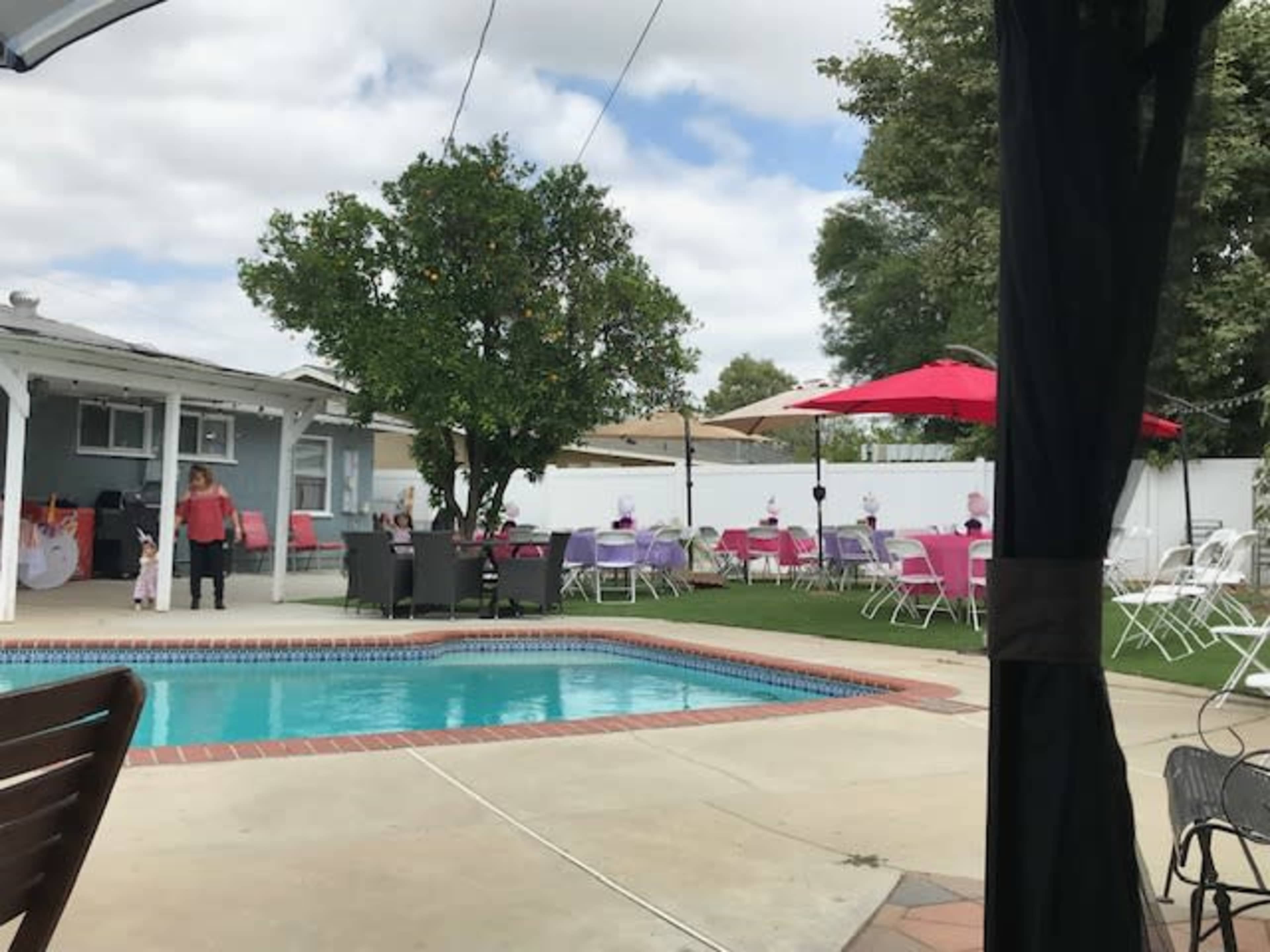 The image shows a backyard pool area with white patio furniture, red umbrellas, and tables set up for an outdoor event.