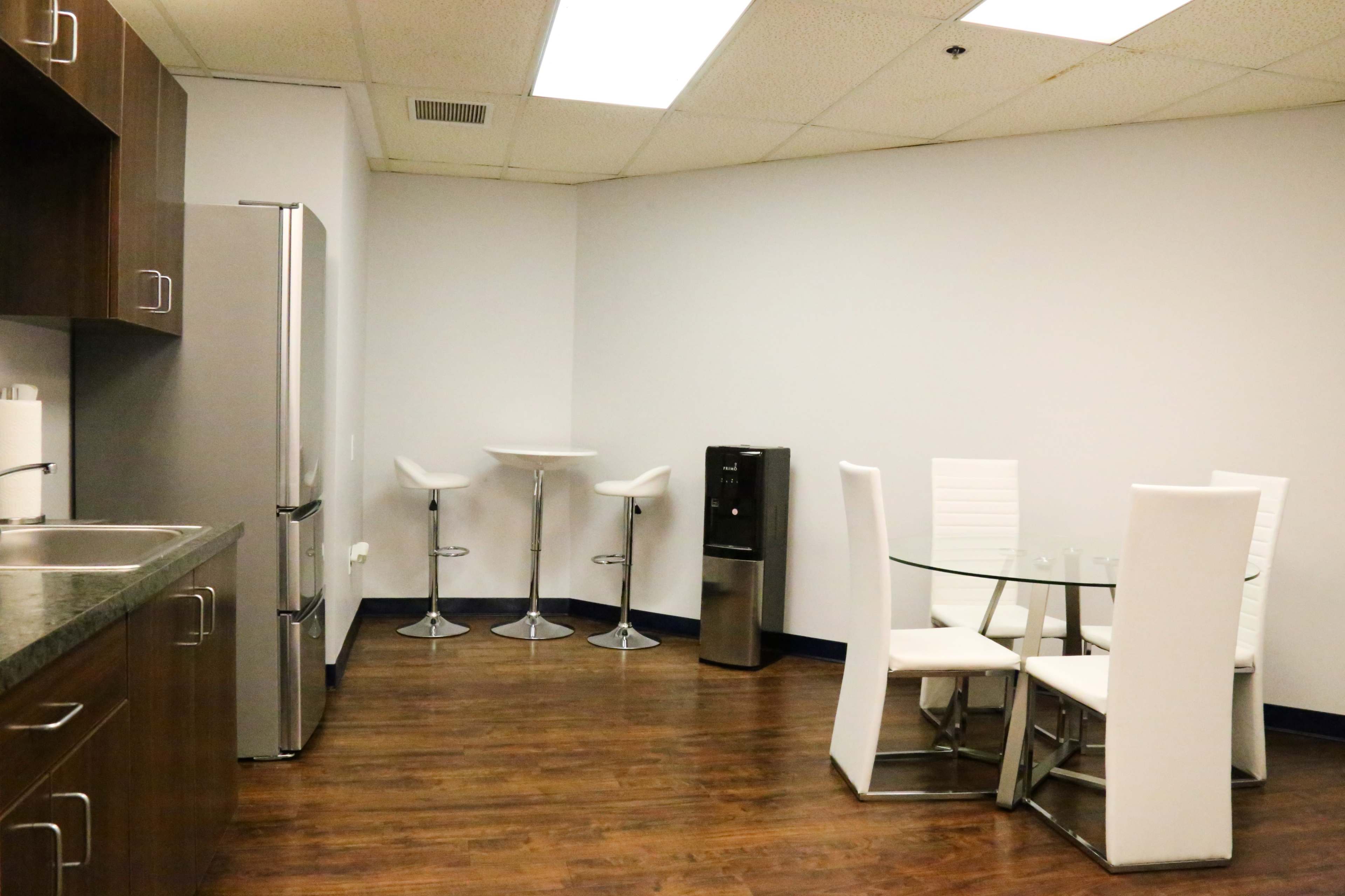 A modern kitchen area featuring a stainless steel refrigerator, a glass dining table with white chairs, and a water dispenser on the side.