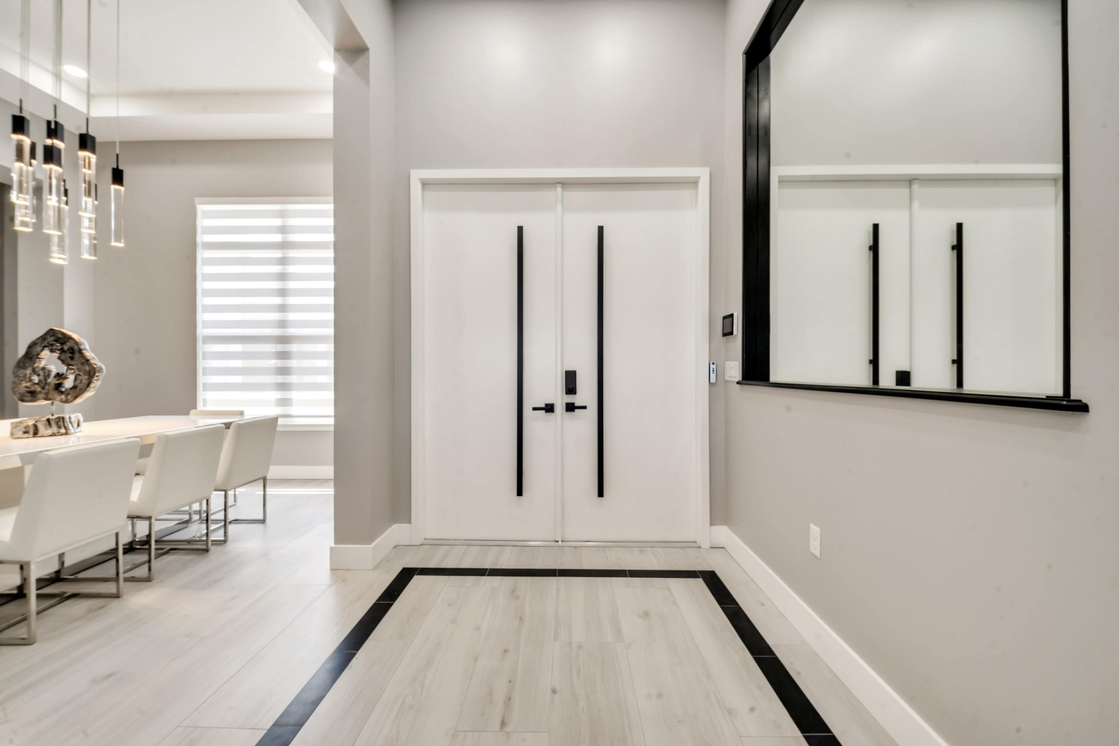 The image shows a modern entrance hallway featuring double white doors, a large mirror, and a view into a dining area with white chairs and a decorative piece on a table.