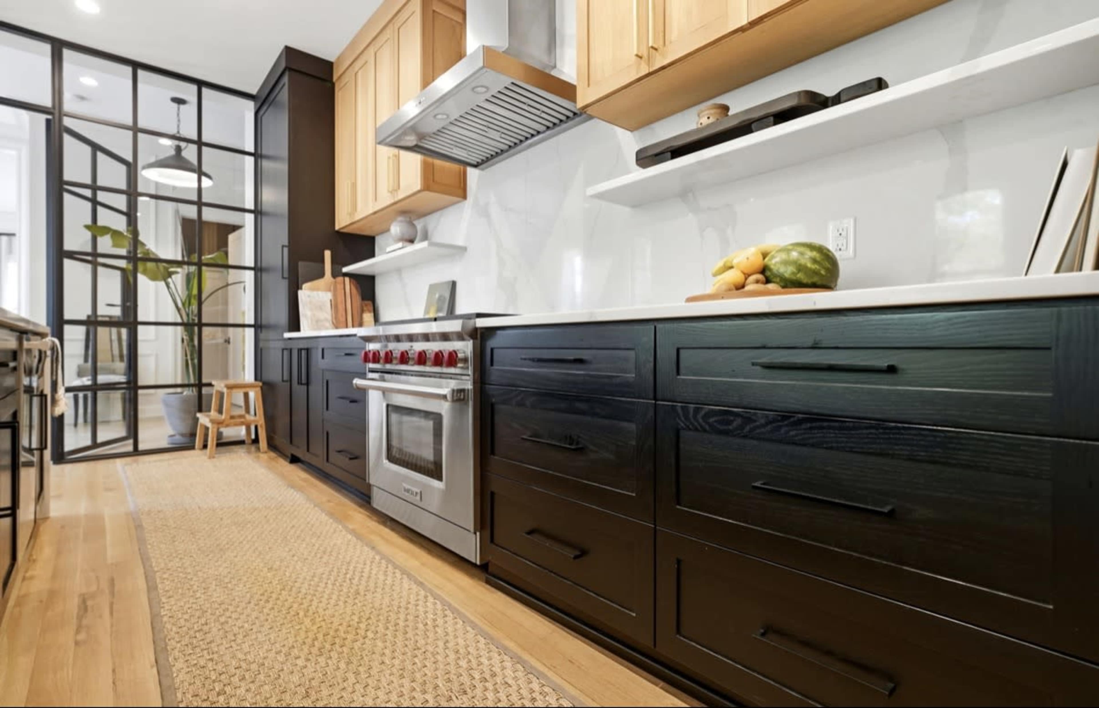 The image shows a modern kitchen with dark and light wood cabinetry, a stainless steel oven, and a marble backsplash, featuring a woven rug on the floor.