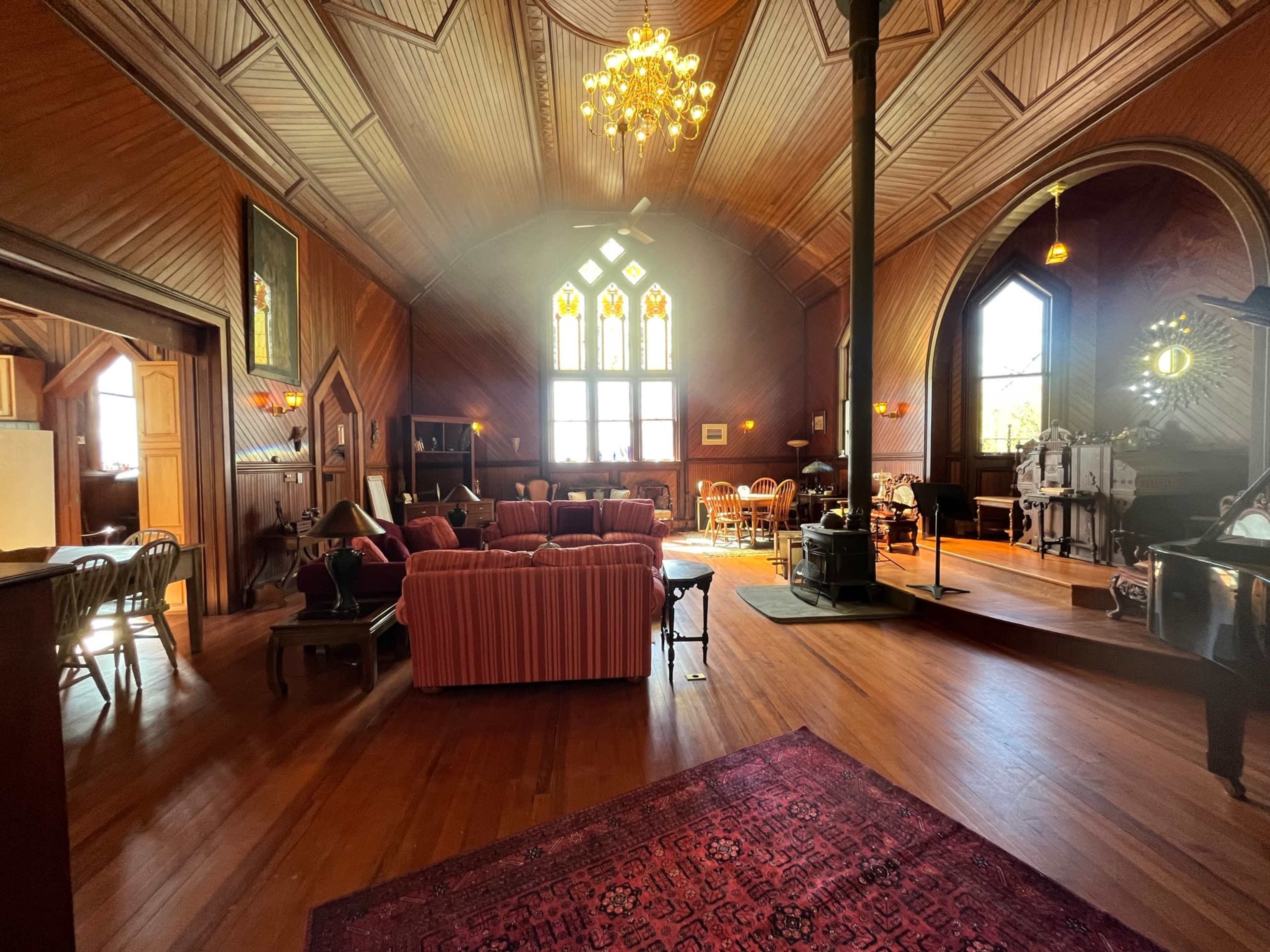A large, wooden-paneled room with a high ceiling, featuring a central seating area, a dining table, and a decorative stove.