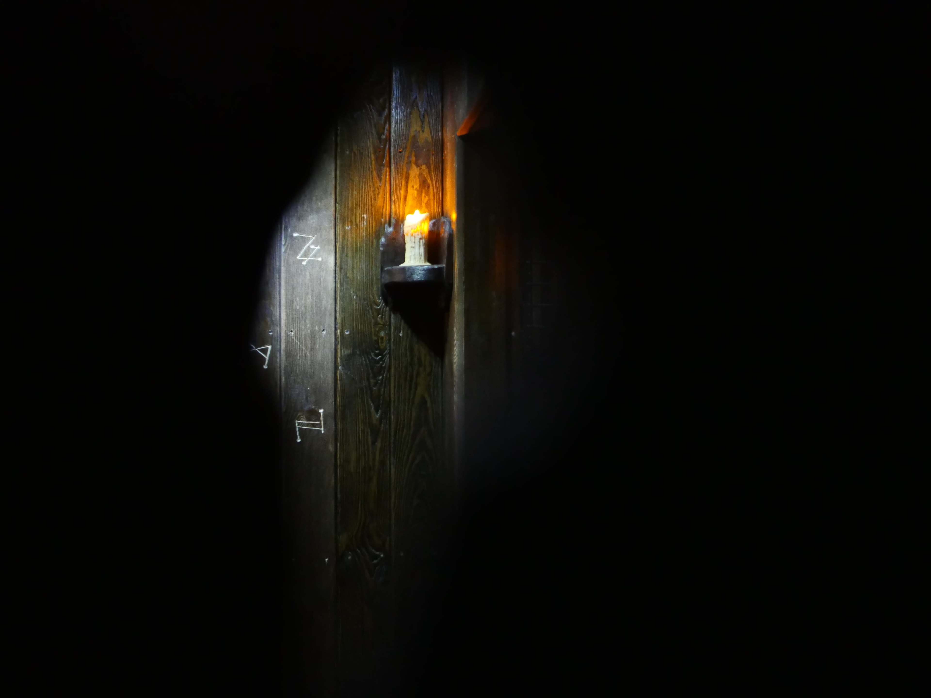 A candle illuminates a wooden wall, partially obscured by a dark opening.