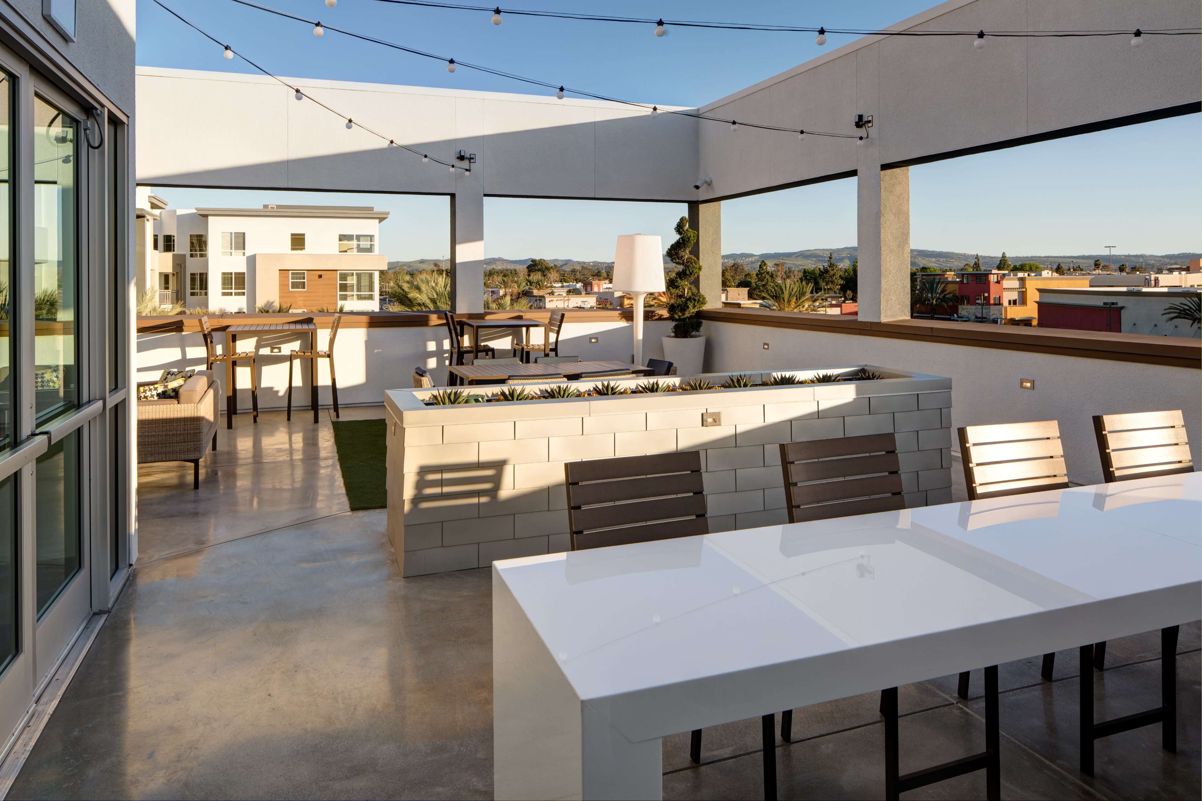 The image shows a modern rooftop patio with a dining table, seating areas, and string lights, overlooking a cityscape.
