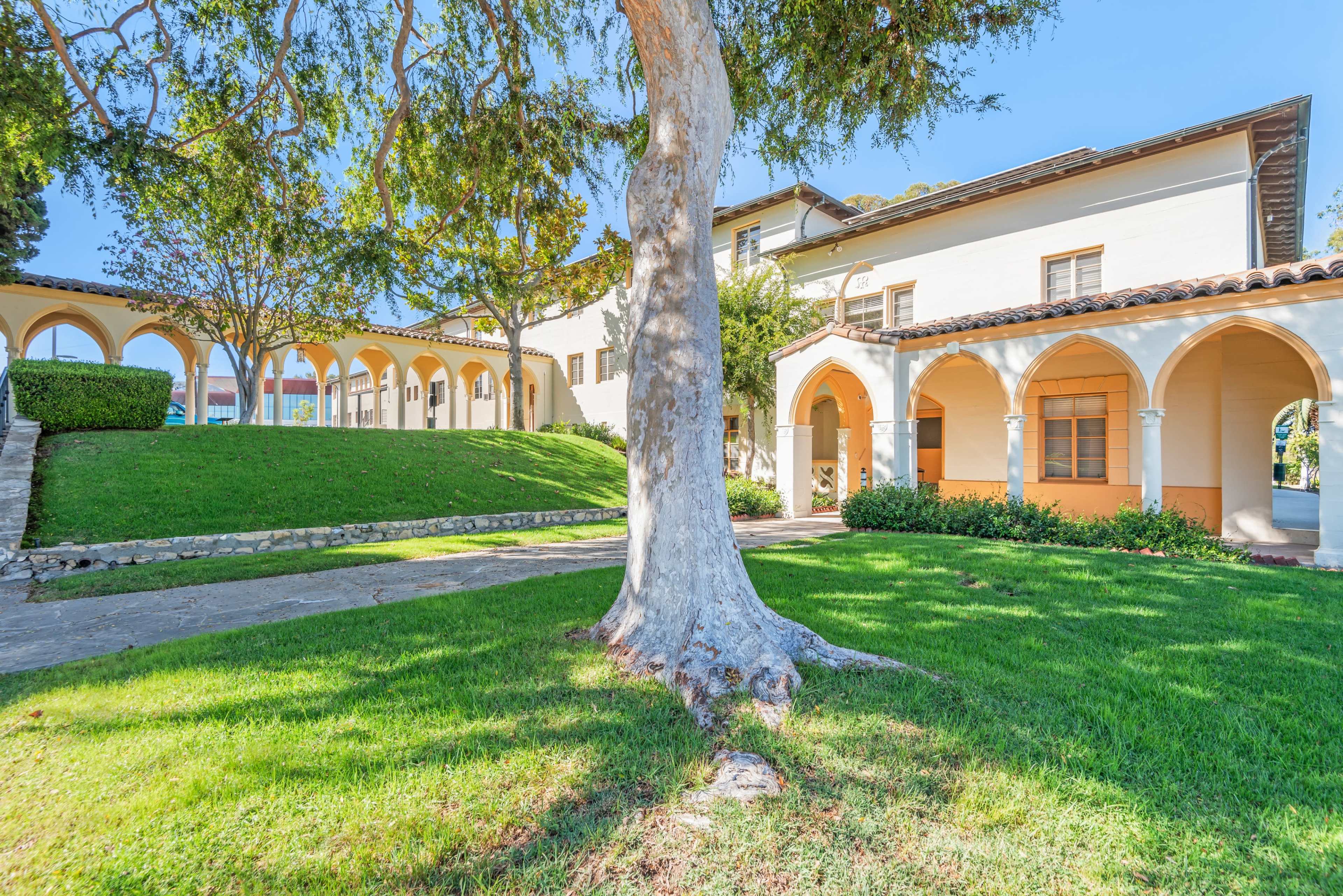 The image shows a well-maintained building with arched entrances and a large tree in a grassy courtyard.