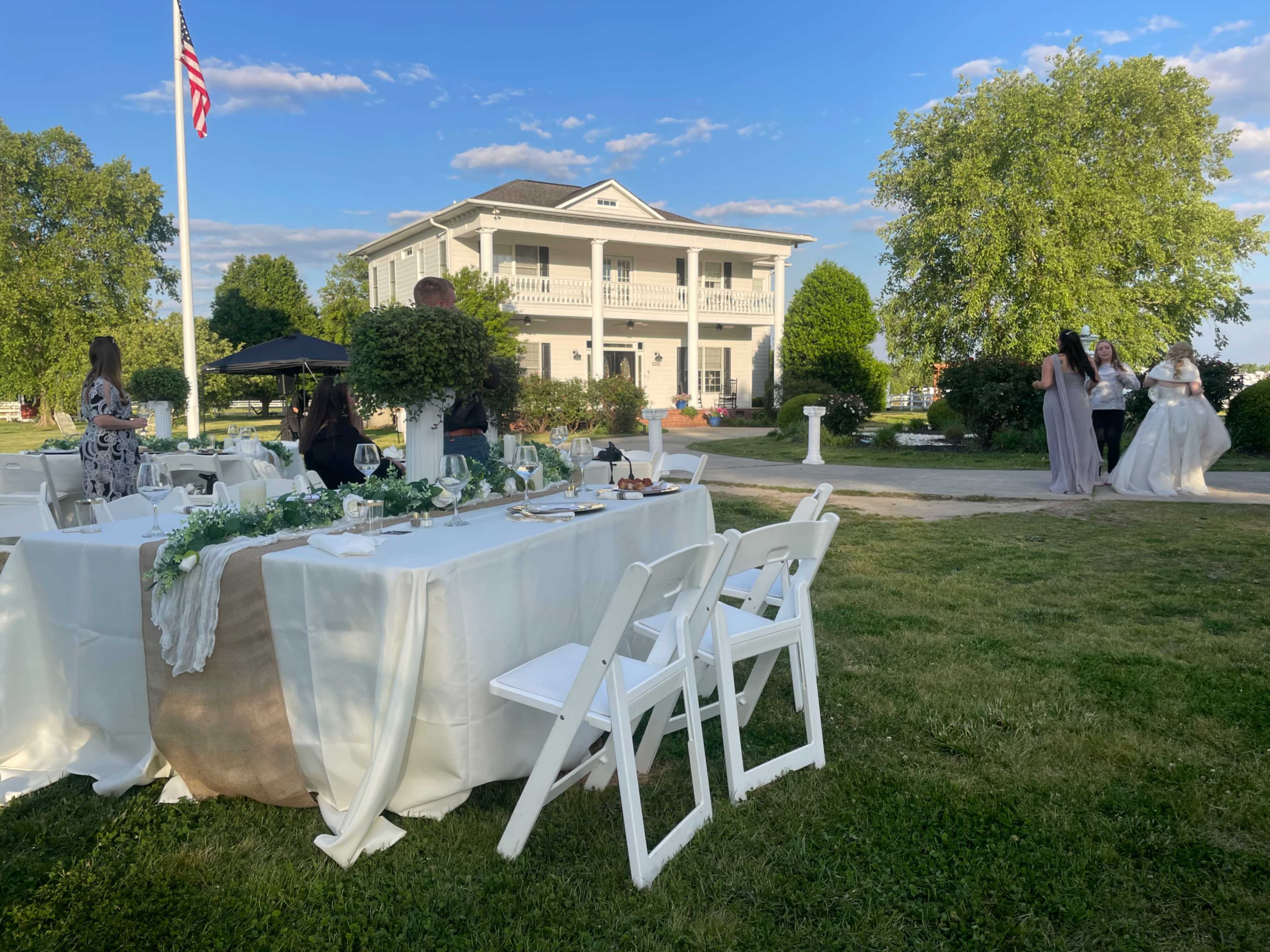 A wedding reception is set up outdoors with tables draped in white cloth, while guests mingle in front of a large, two-story venue surrounded by greenery.