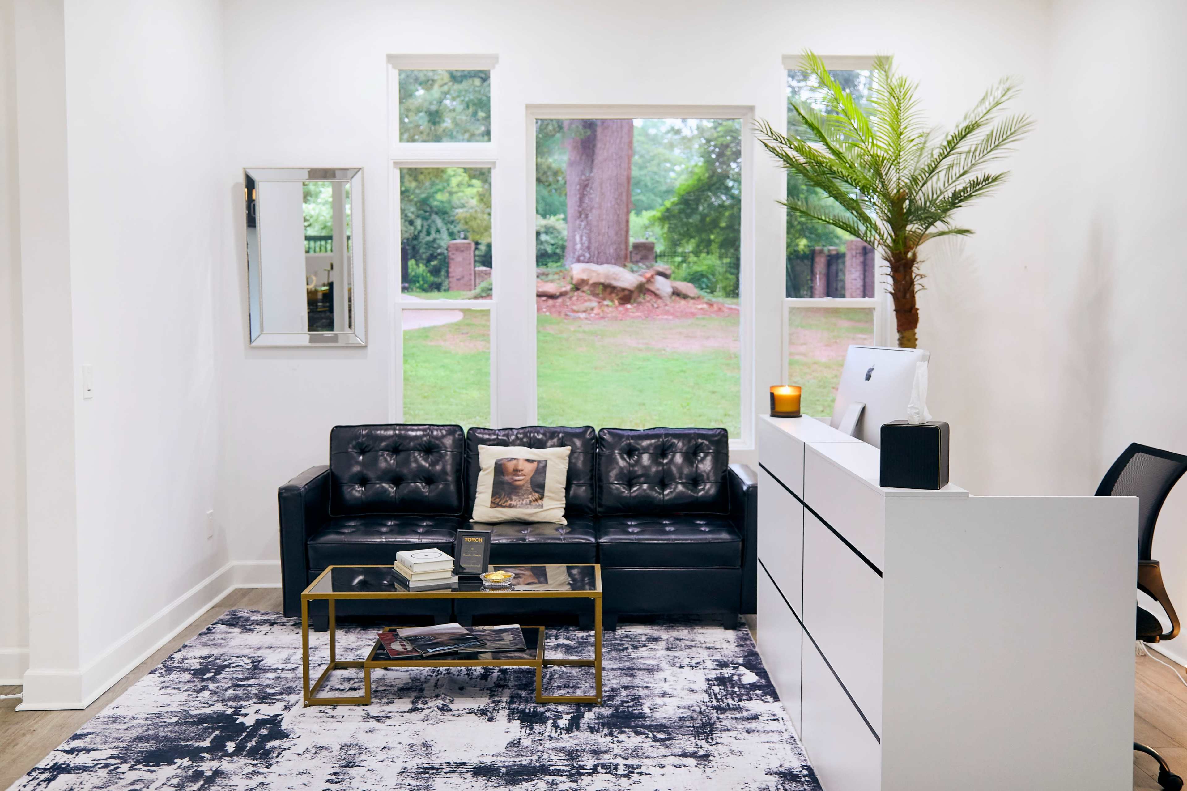 The image shows a modern living room with a black leather sofa, a glass-top coffee table, and large windows overlooking a green outdoor space.