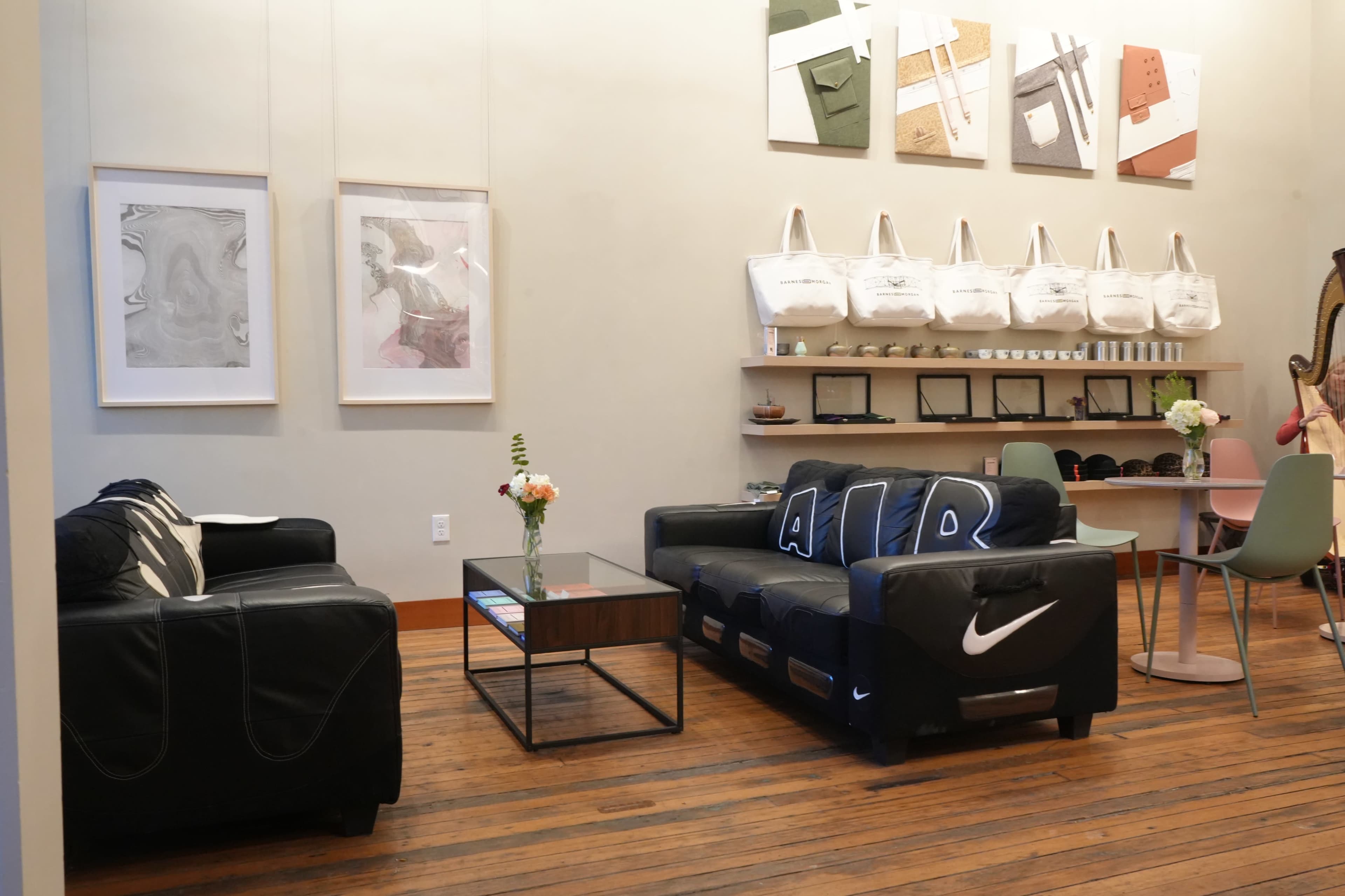 The image shows a modern lounge area with black sofas, a glass coffee table, artwork on the walls, and a display of tote bags and chairs nearby.
