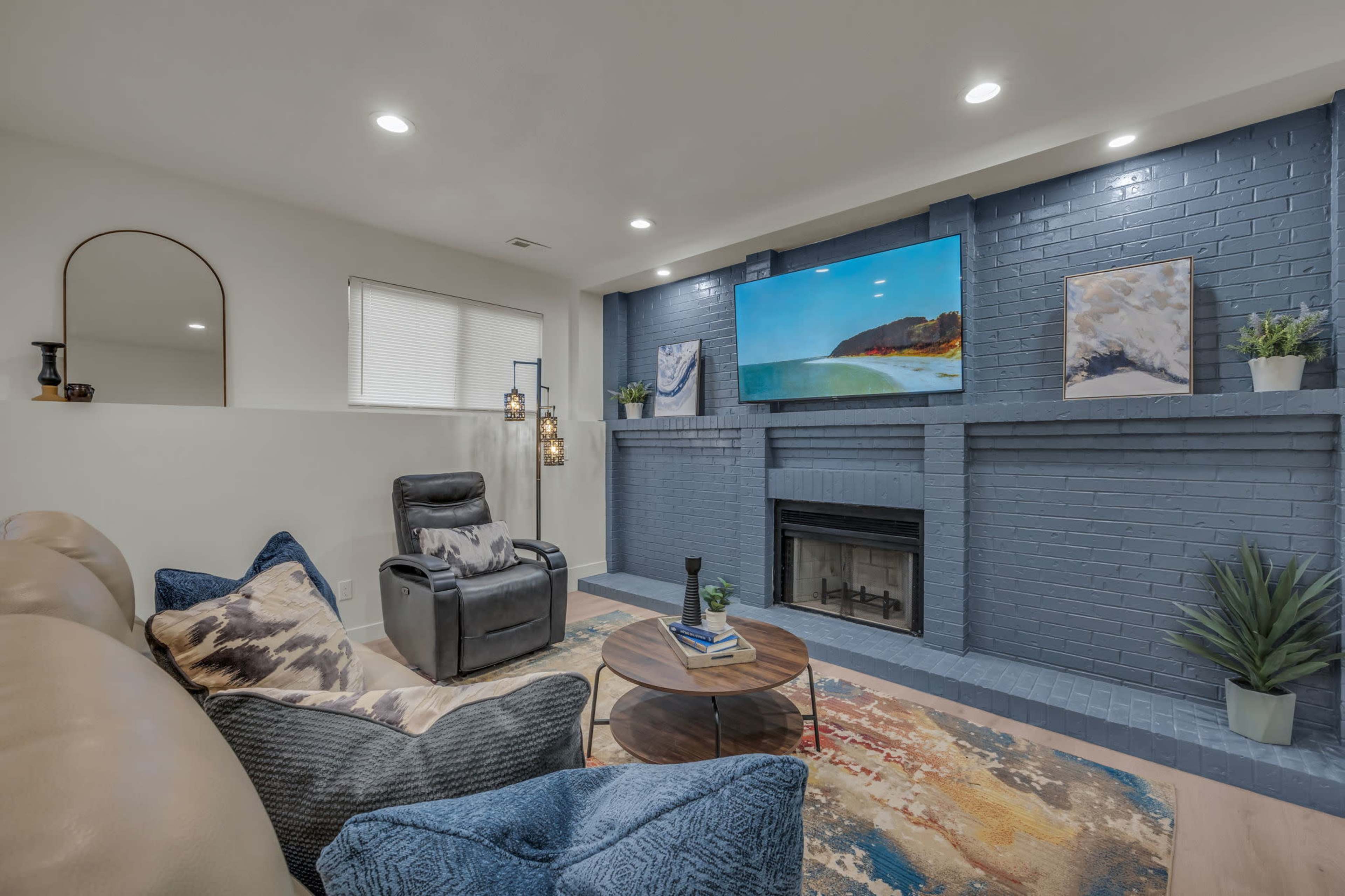 A cozy living room features a fireplace surrounded by a blue brick wall, a large TV mounted above, and comfortable seating with a coffee table and decorative elements.
