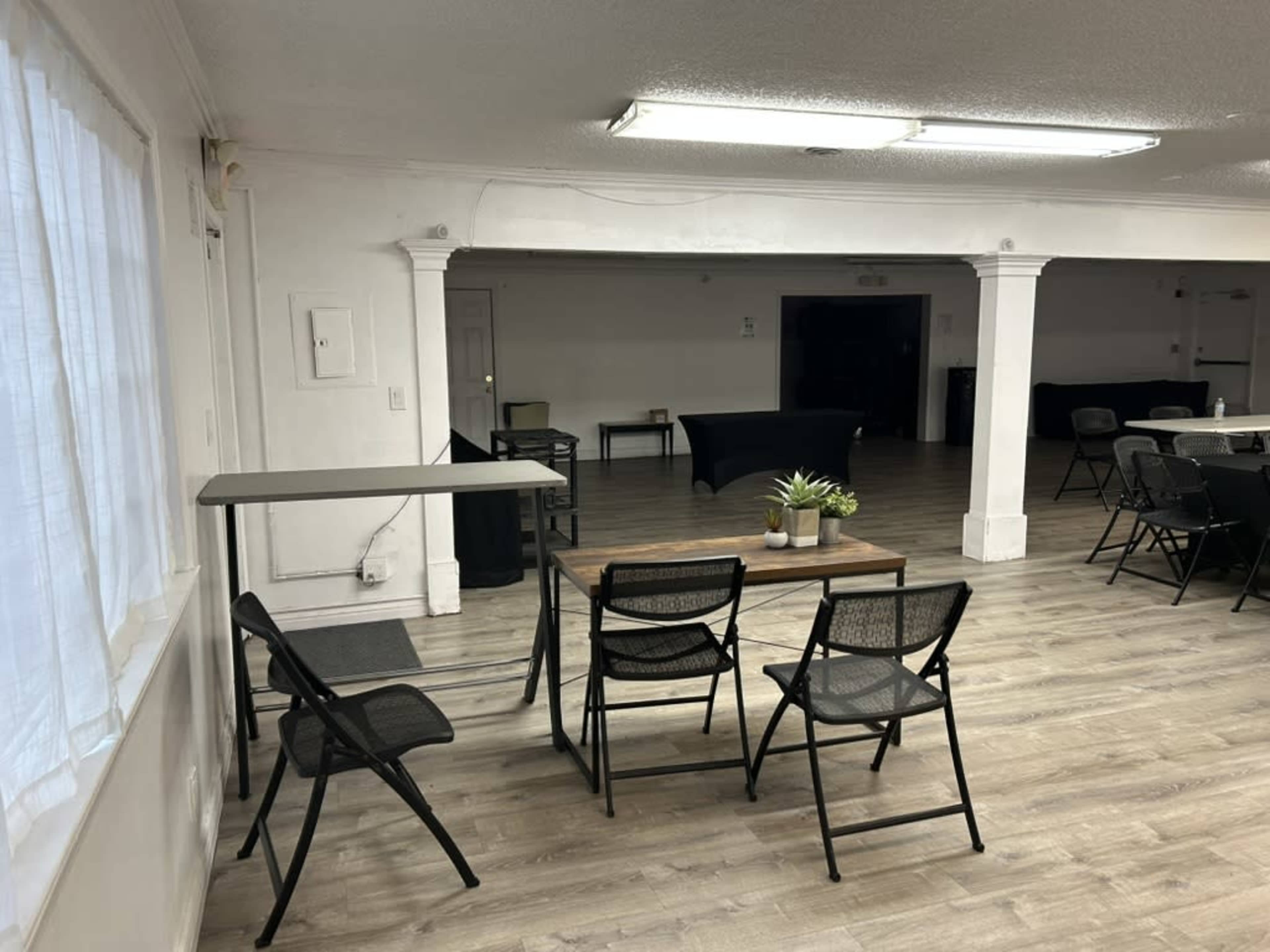 The image shows a spacious room with wooden flooring, featuring a table with two chairs, a plant, and several empty tables and chairs arranged for an event.