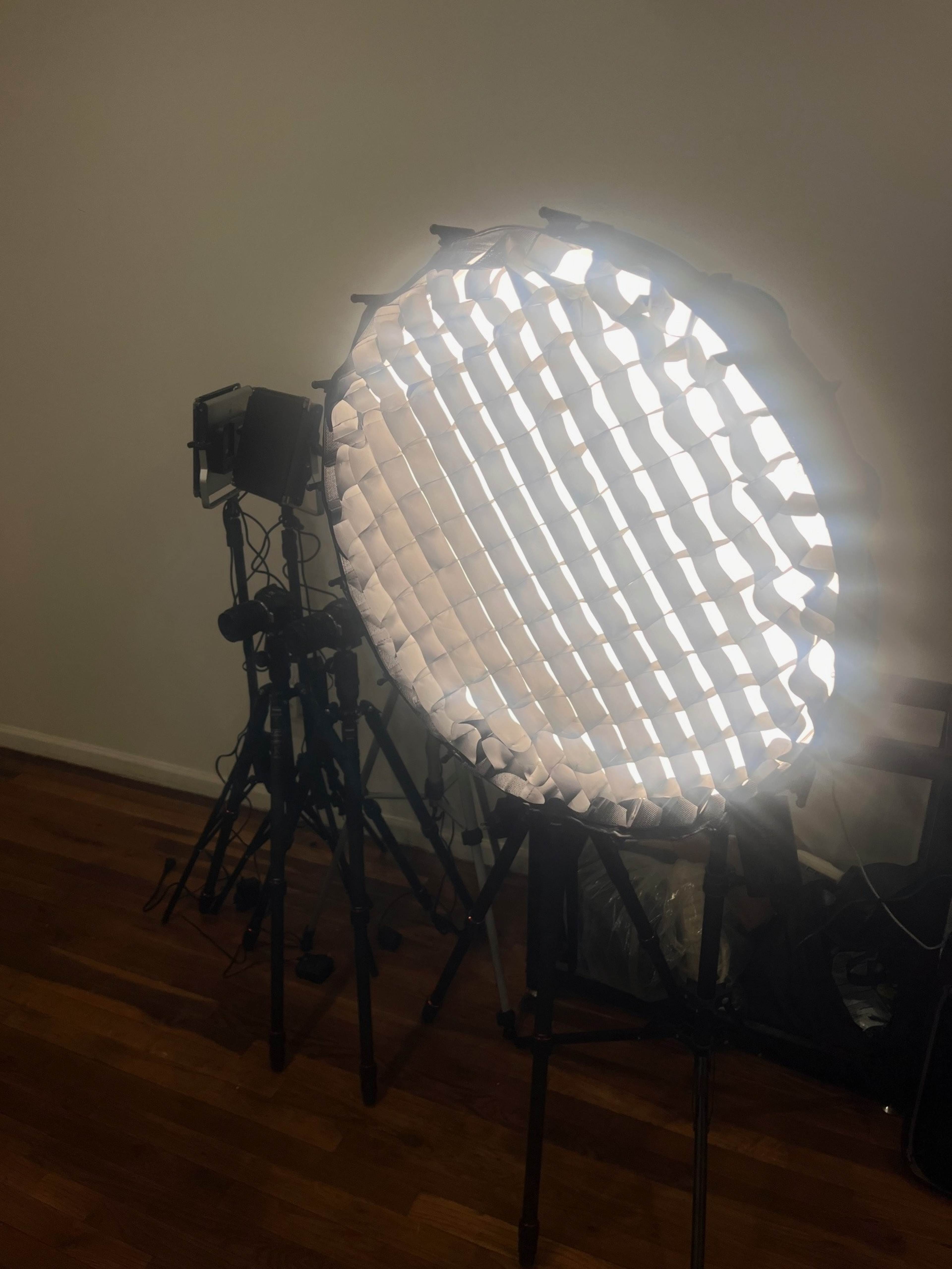 The image shows a round softbox with a grid attached, positioned next to a studio light on a tripod, set against a plain wall and wooden floor.