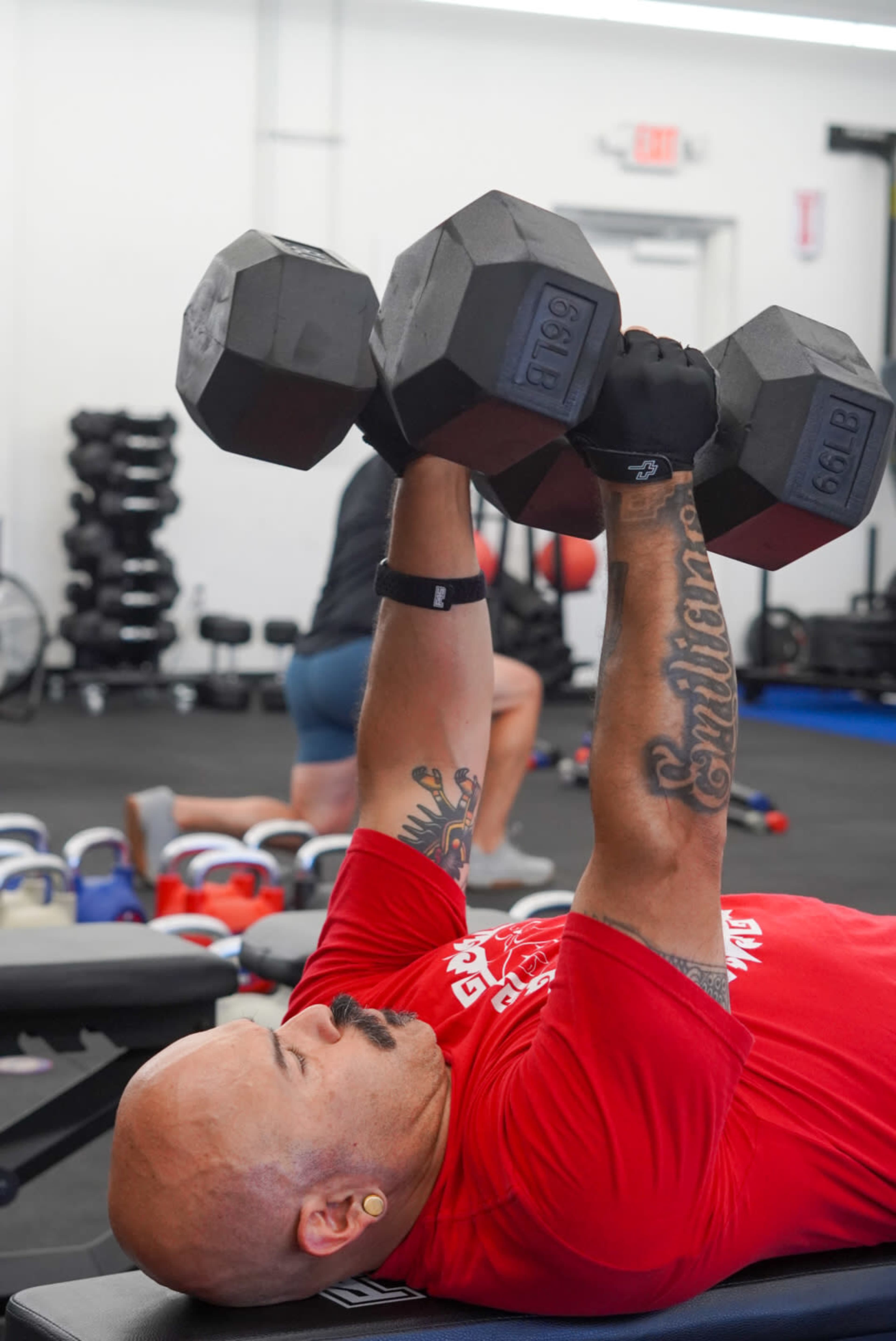 A person lies on a bench in a gym, lifting dumbbells overhead with one arm while others exercise in the background.