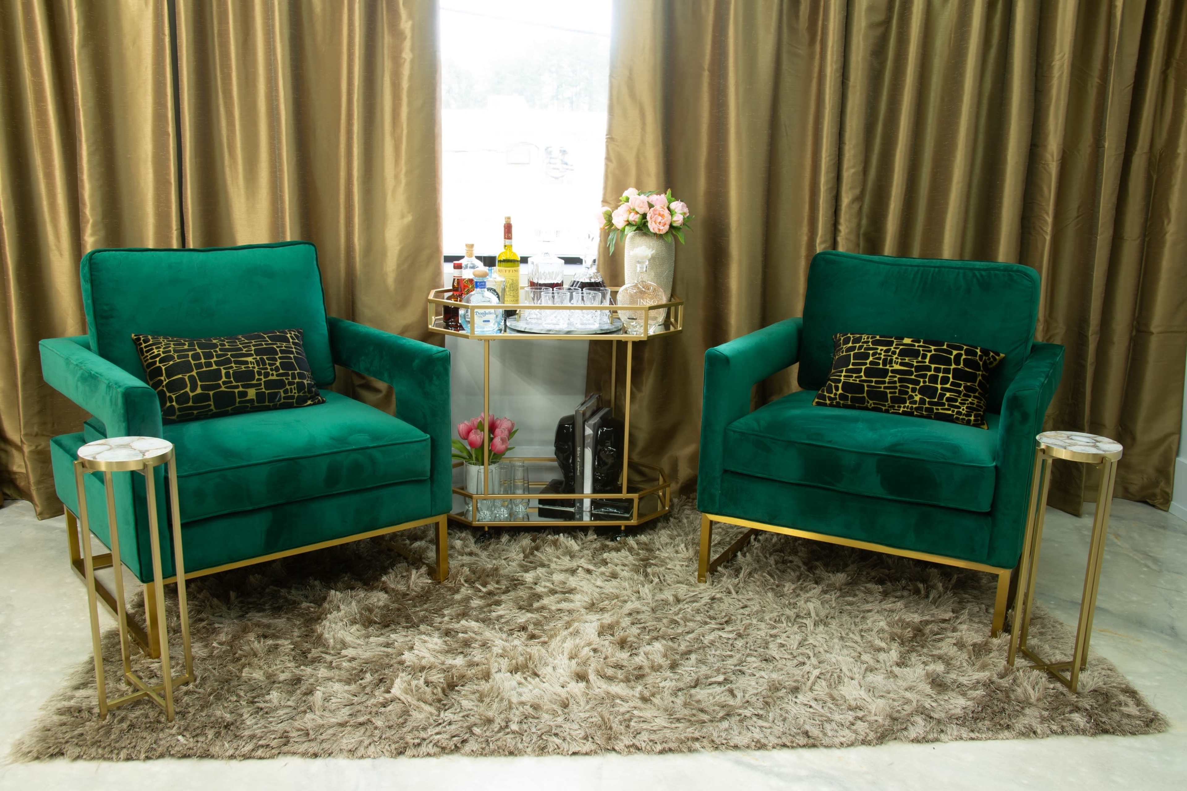 Two green velvet armchairs with gold accents flanking a glass-top side table on a plush area rug in a room with golden curtains.
