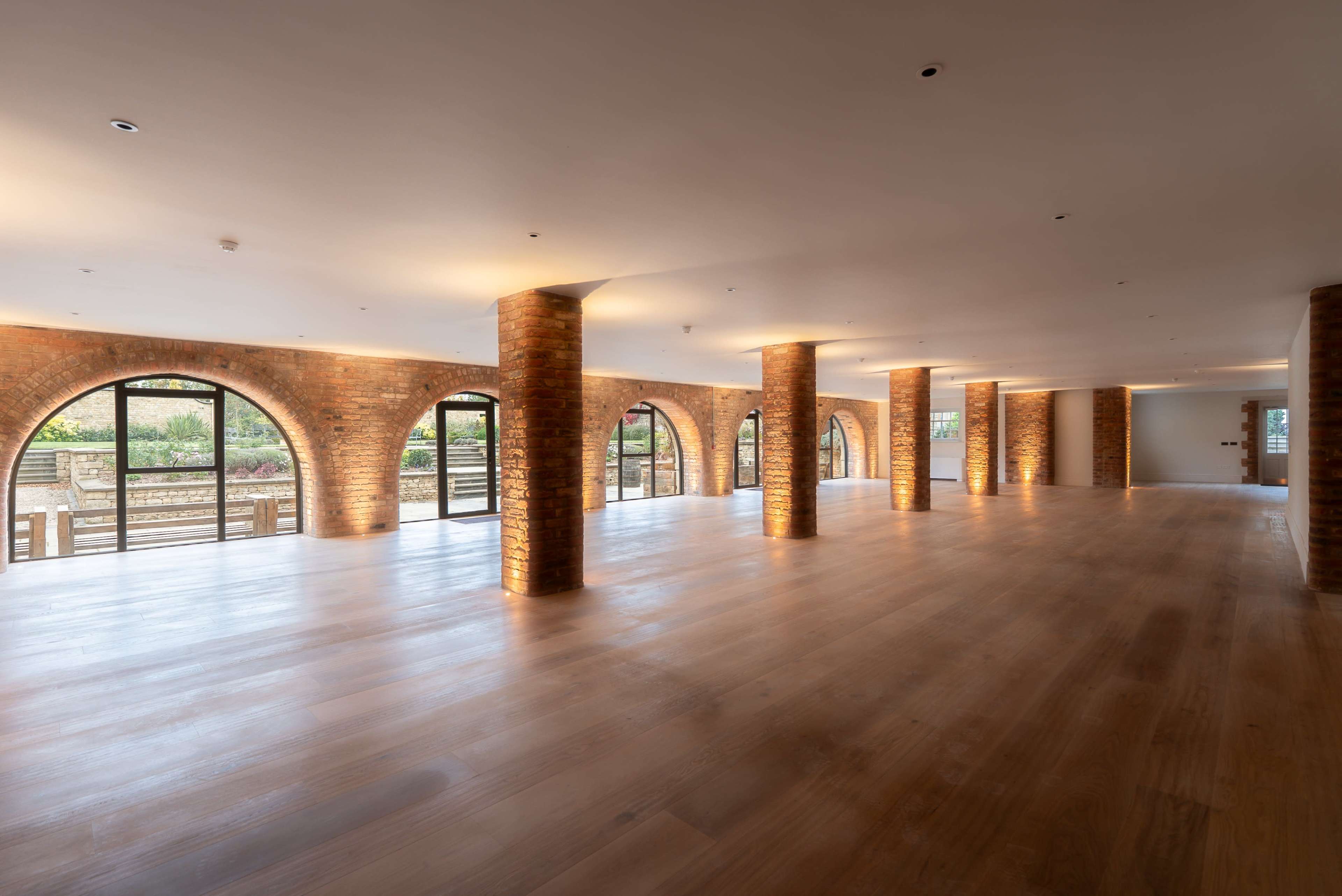 The image shows a large, open room with brick arches and wooden flooring, featuring large windows that provide natural light and a view of an outdoor space.