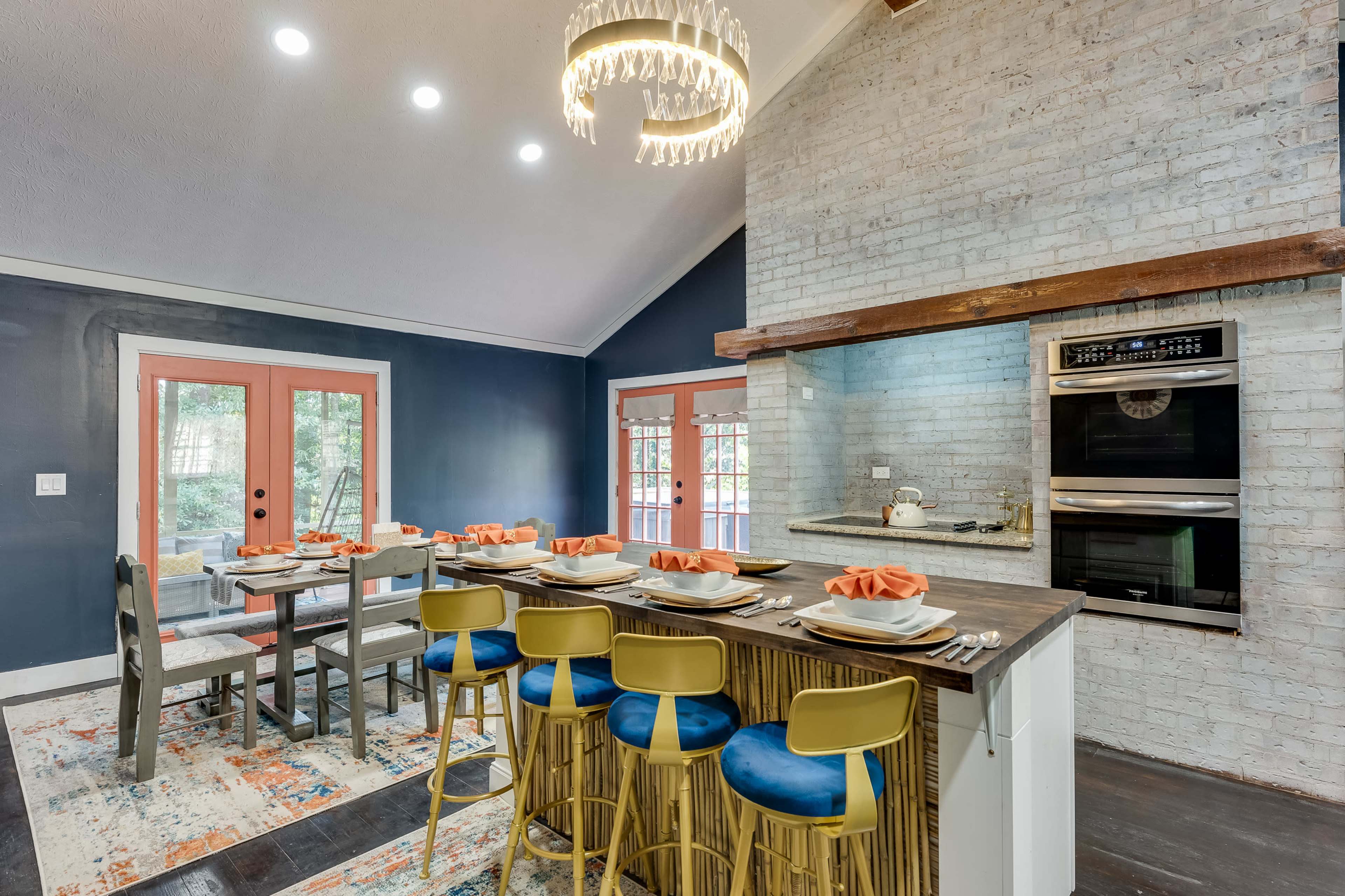 A modern kitchen features a large dining table with orange napkins and a countertop bar with blue stools, set against a backdrop of exposed brick and dark walls.