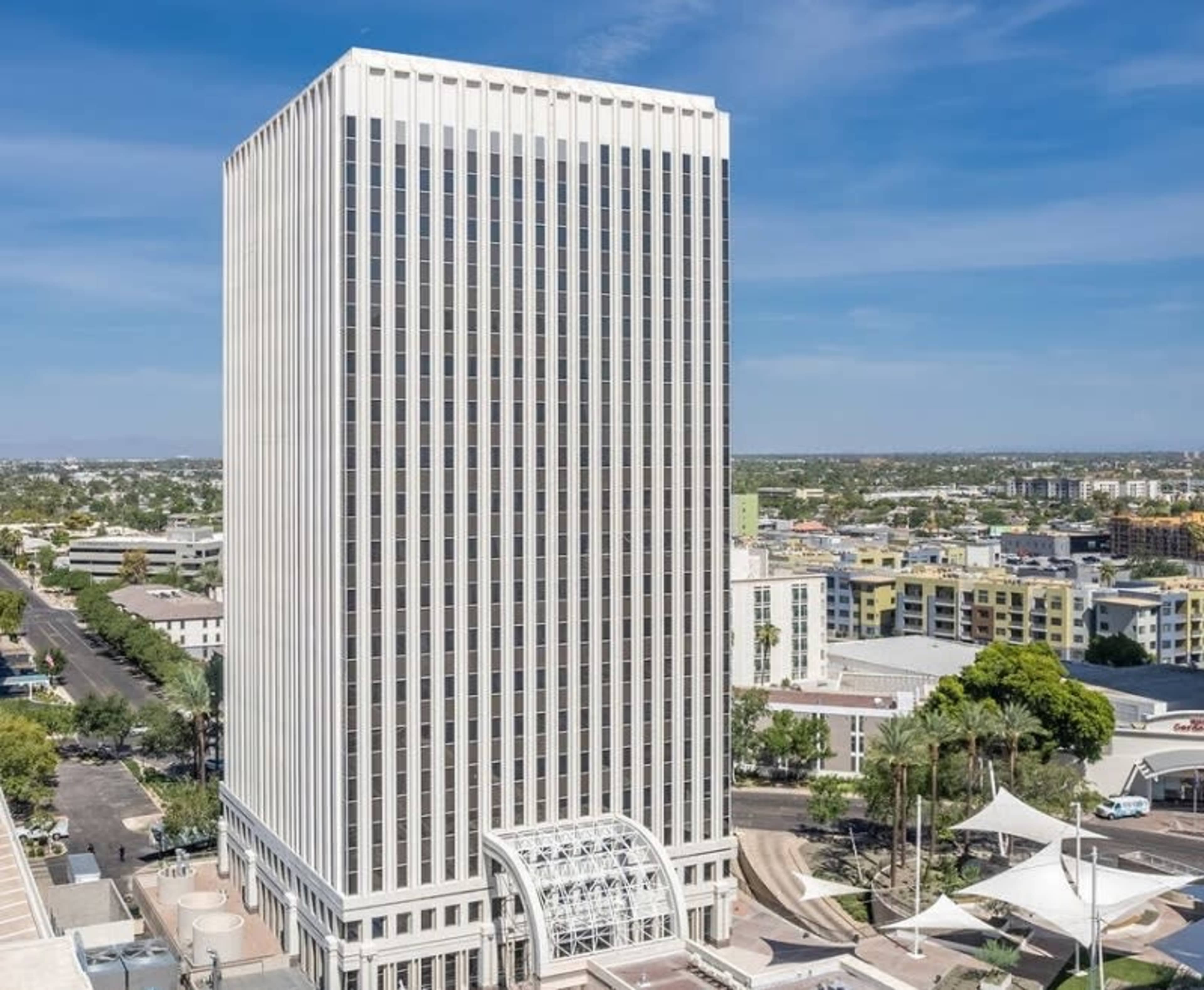 The image shows a tall, modern office building with a white facade and vertical lines, set against a clear blue sky and a city landscape in the background.