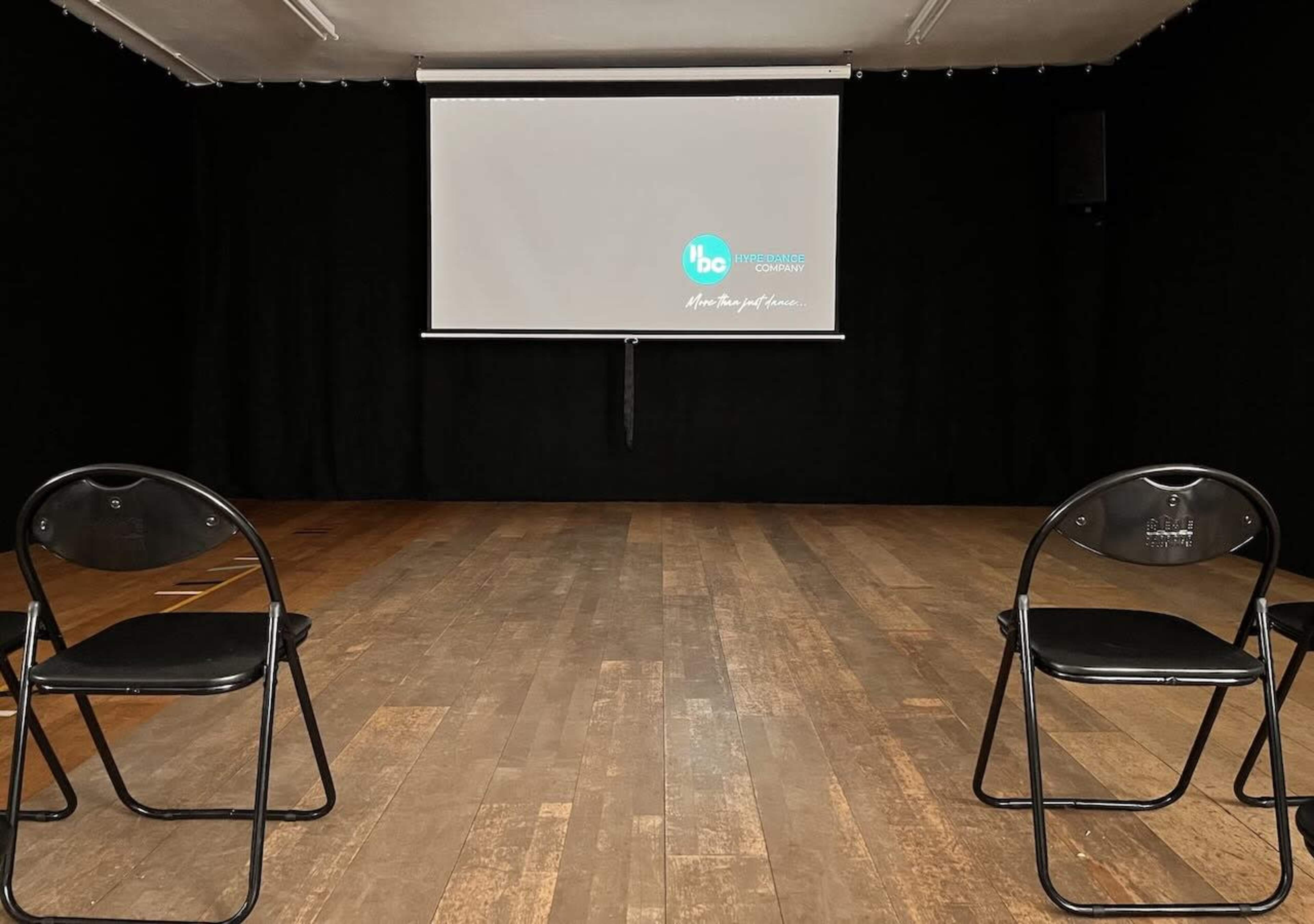 The image shows a dimly lit stage area with two black folding chairs facing a blank projection screen on a wall.