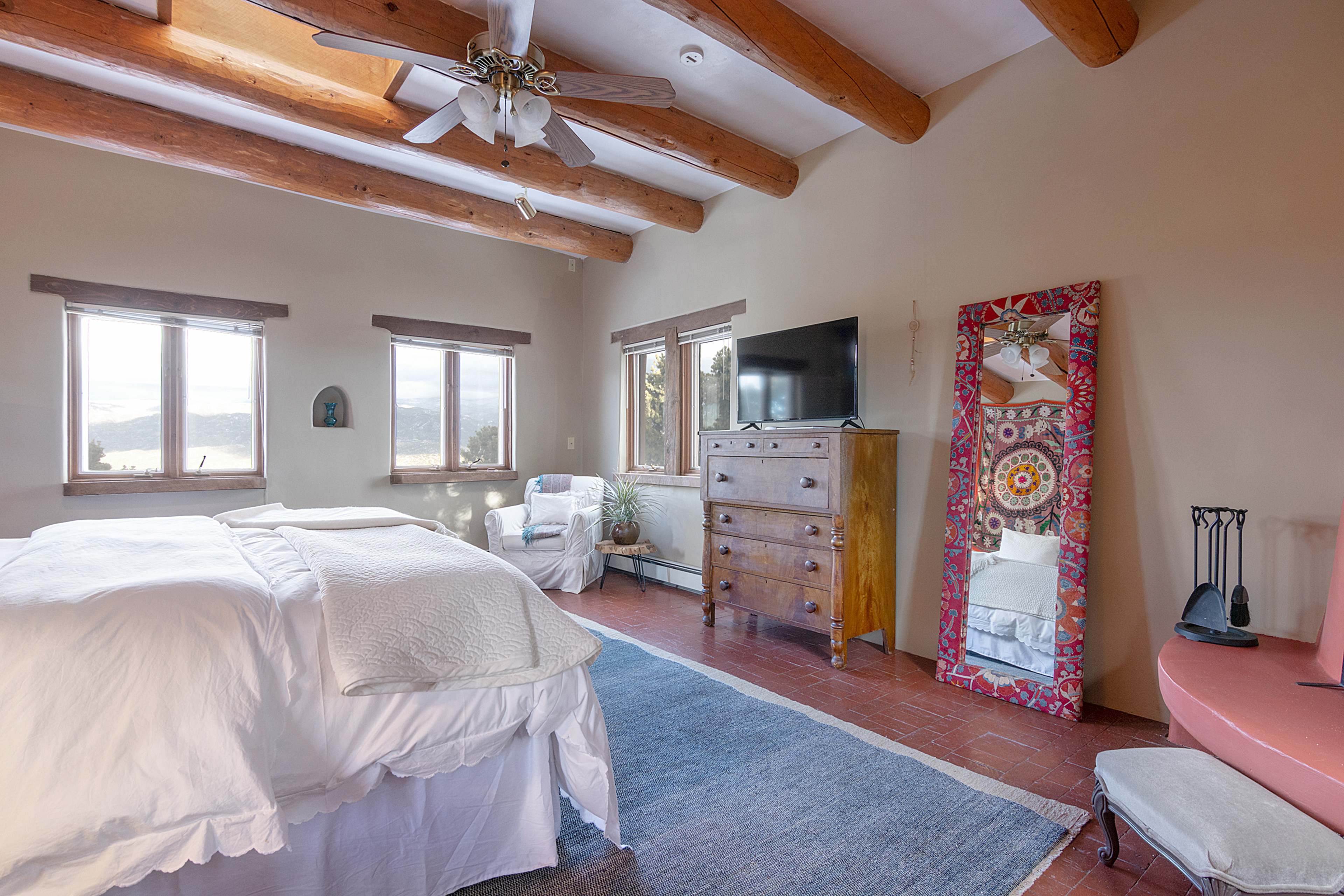 A bedroom features wooden beams, a large bed with white bedding, a dresser, a TV, and a colorful mirror, with windows offering views of the outdoors.