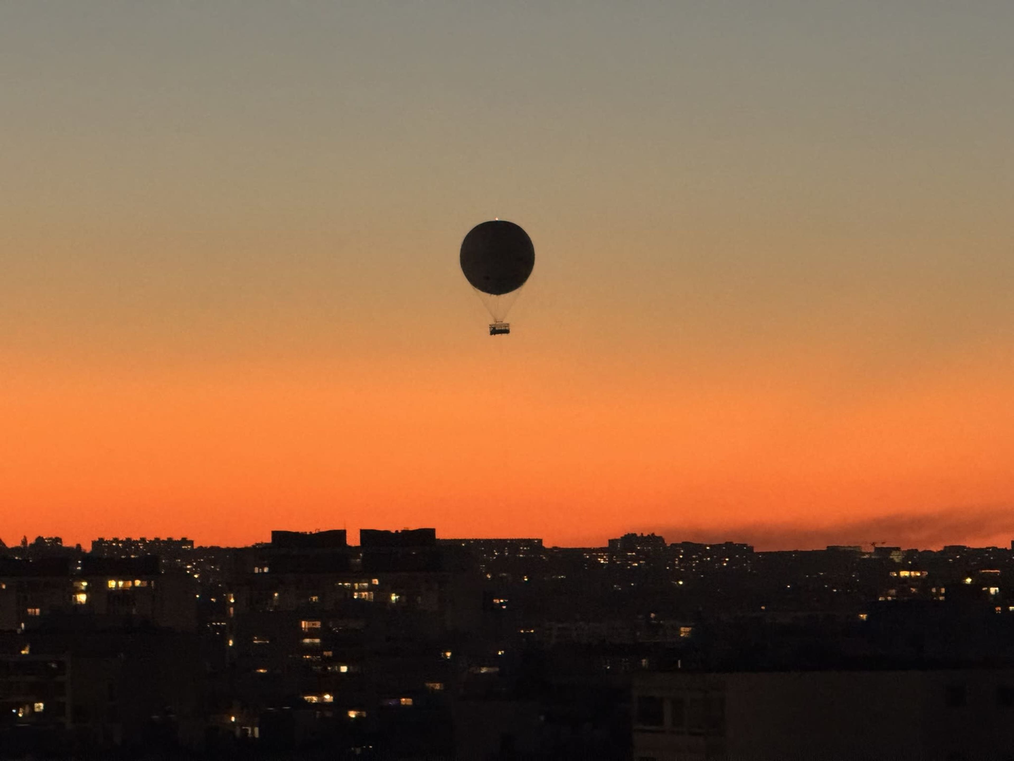 A hot air balloon floats in the twilight sky above a city skyline as sunset hues paint the horizon.
