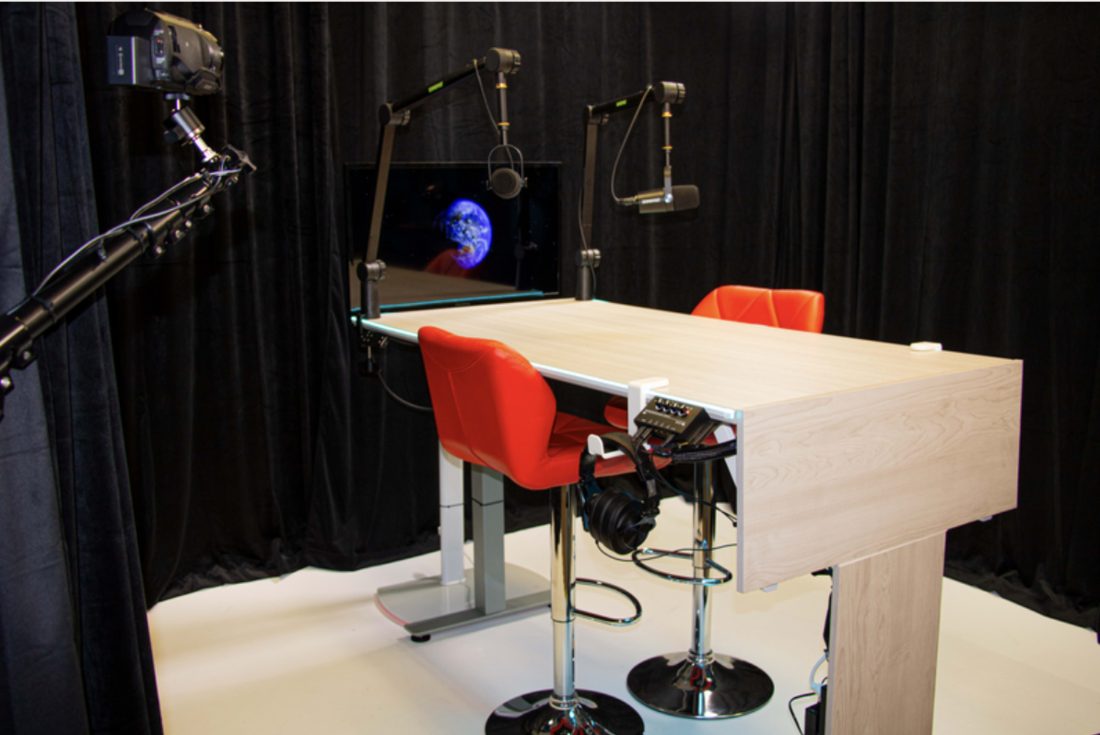 A modern podcasting or broadcasting studio with a wooden table, red chairs, two microphones on adjustable arms, and a large screen displaying an image of Earth.