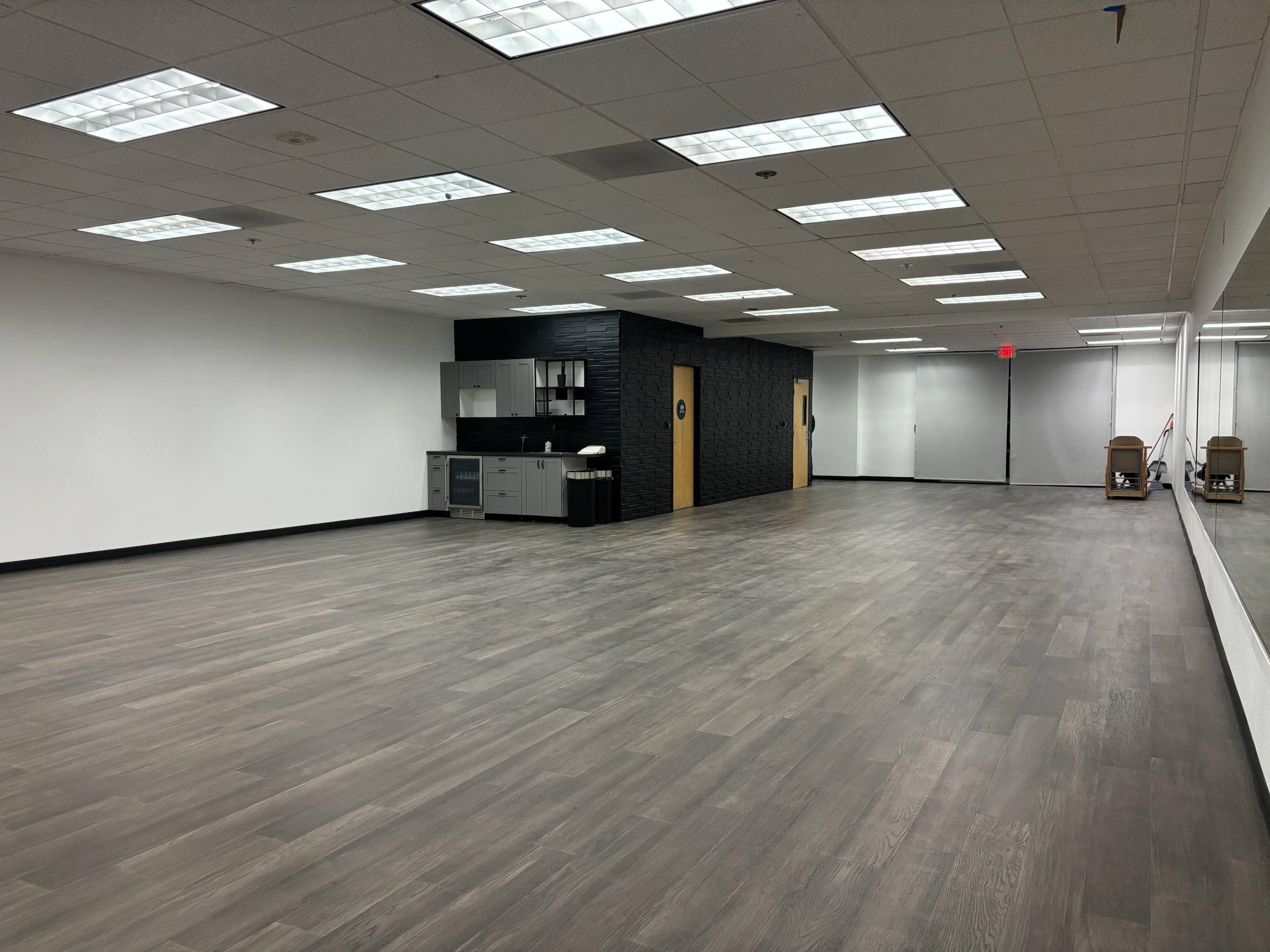 A spacious, empty room with wooden flooring, a kitchenette on one wall, and mirrors along another wall.