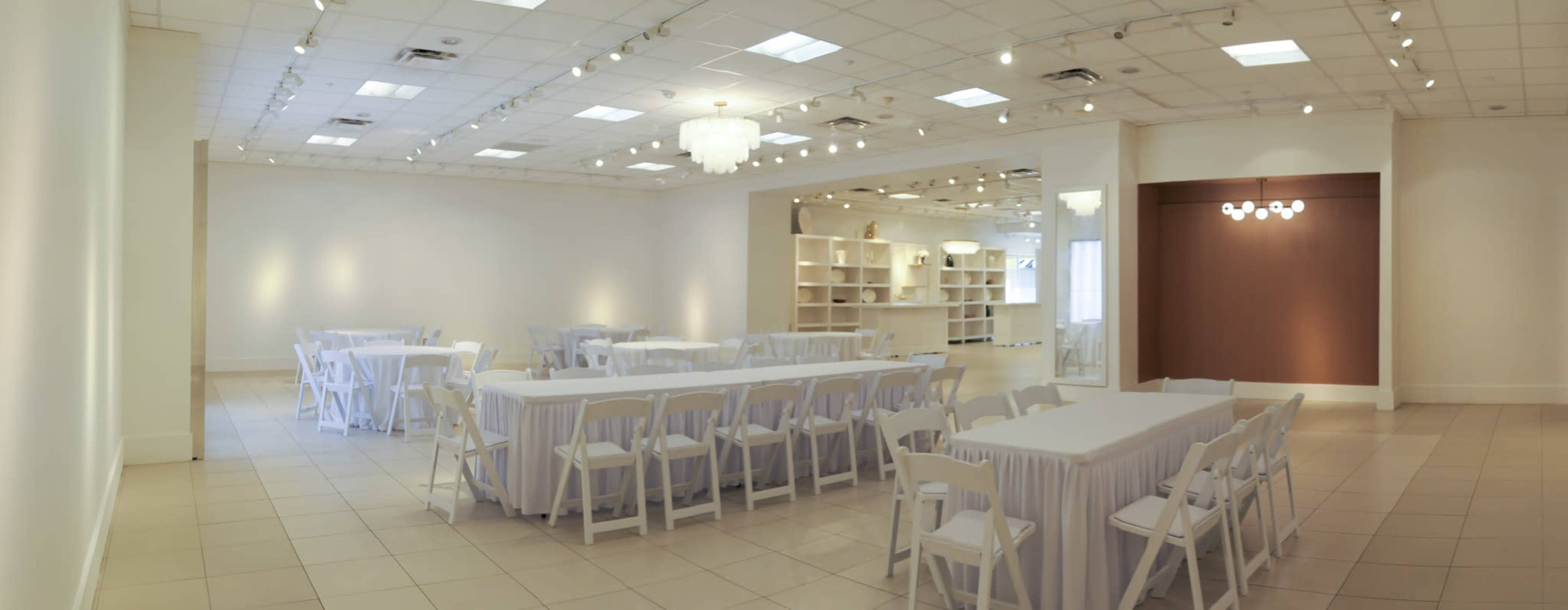 The image shows a spacious event room arranged with several tables covered in white linens and surrounded by white chairs, with soft lighting and a minimalist design.
