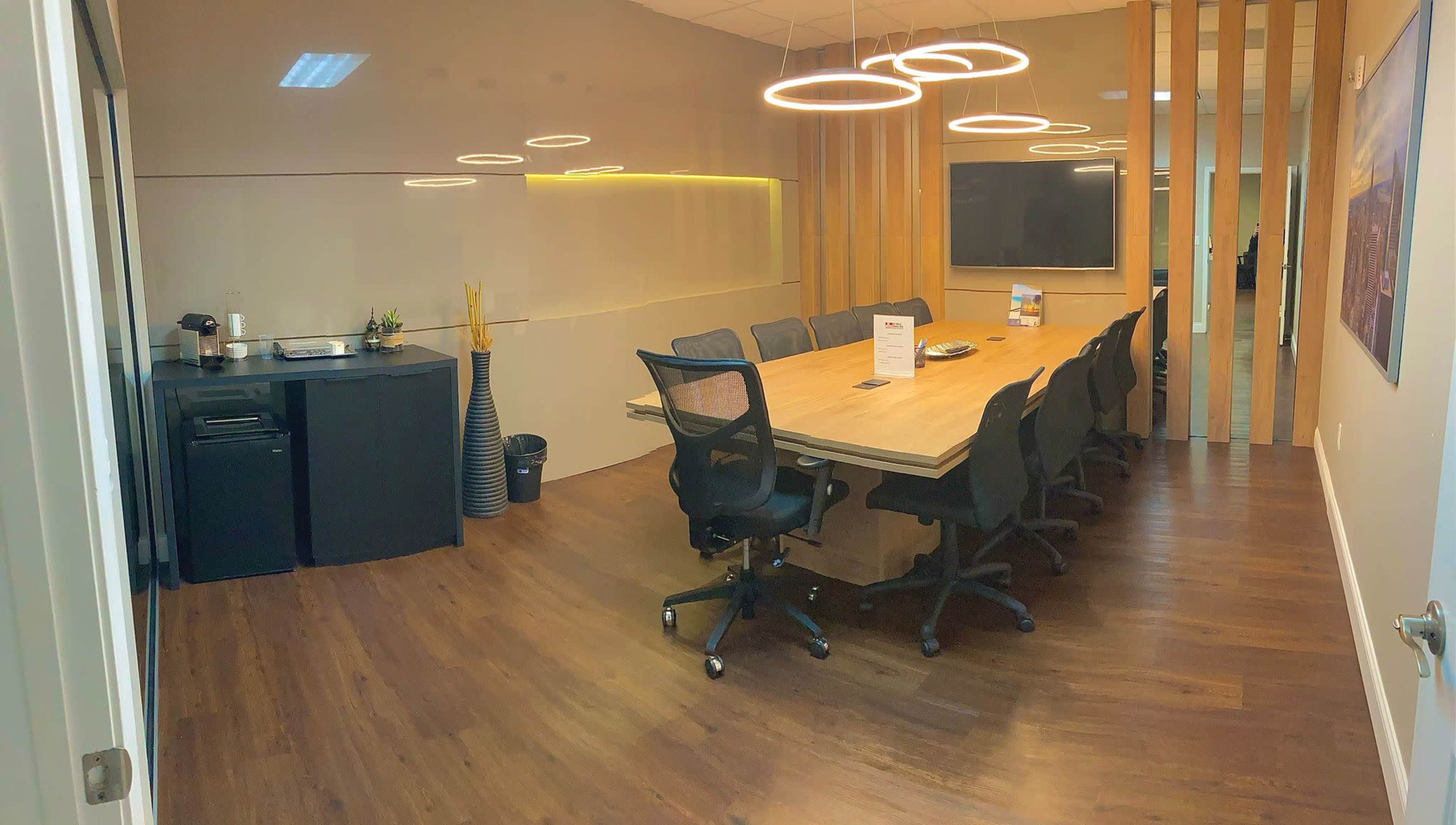 A modern conference room features a large wooden table surrounded by black office chairs, with a coffee station and a sleek cabinet along one wall.