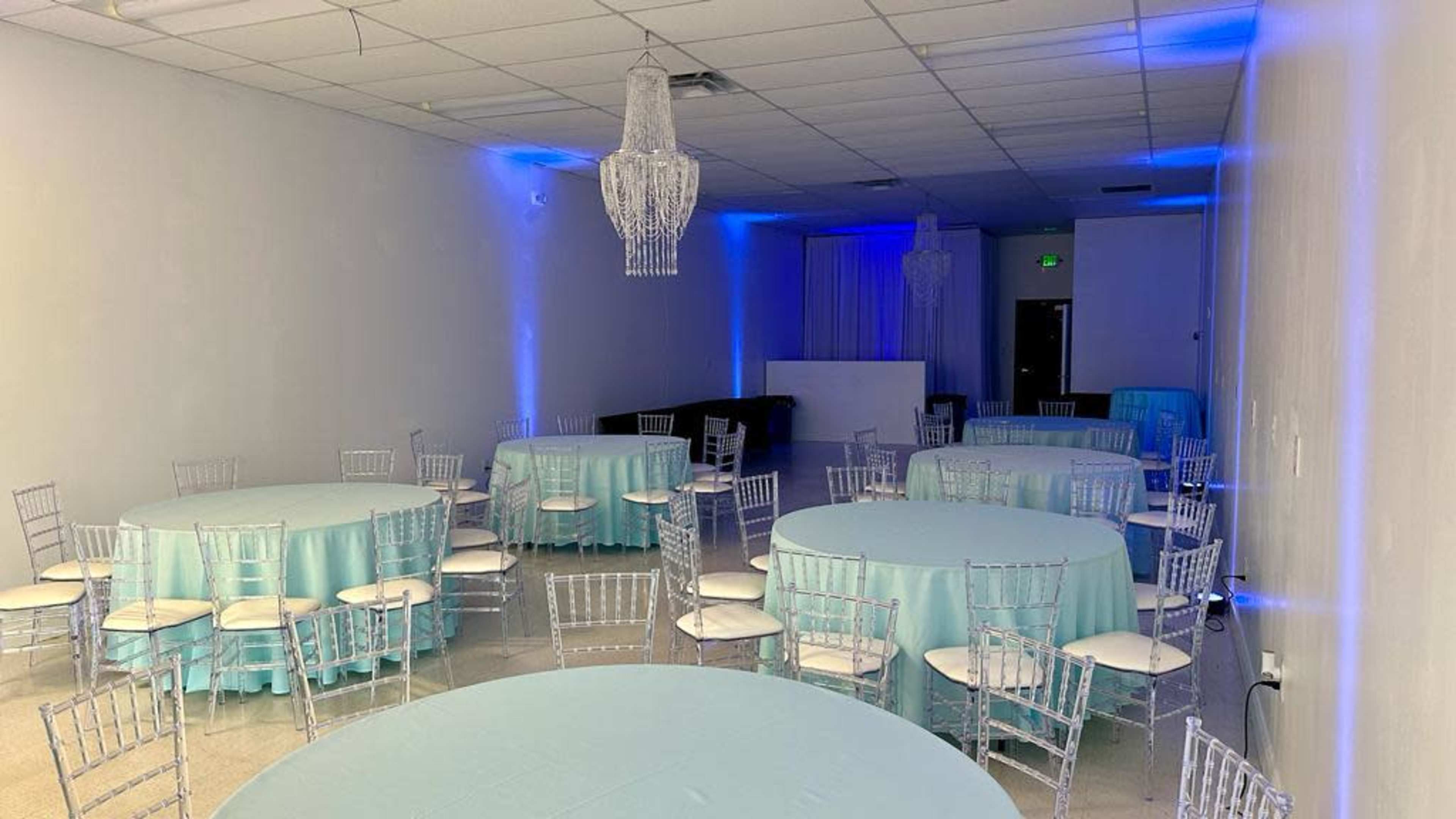 The image shows a spacious event room with several round tables covered in light blue tablecloths, white chairs, and blue lighting accents on the walls.