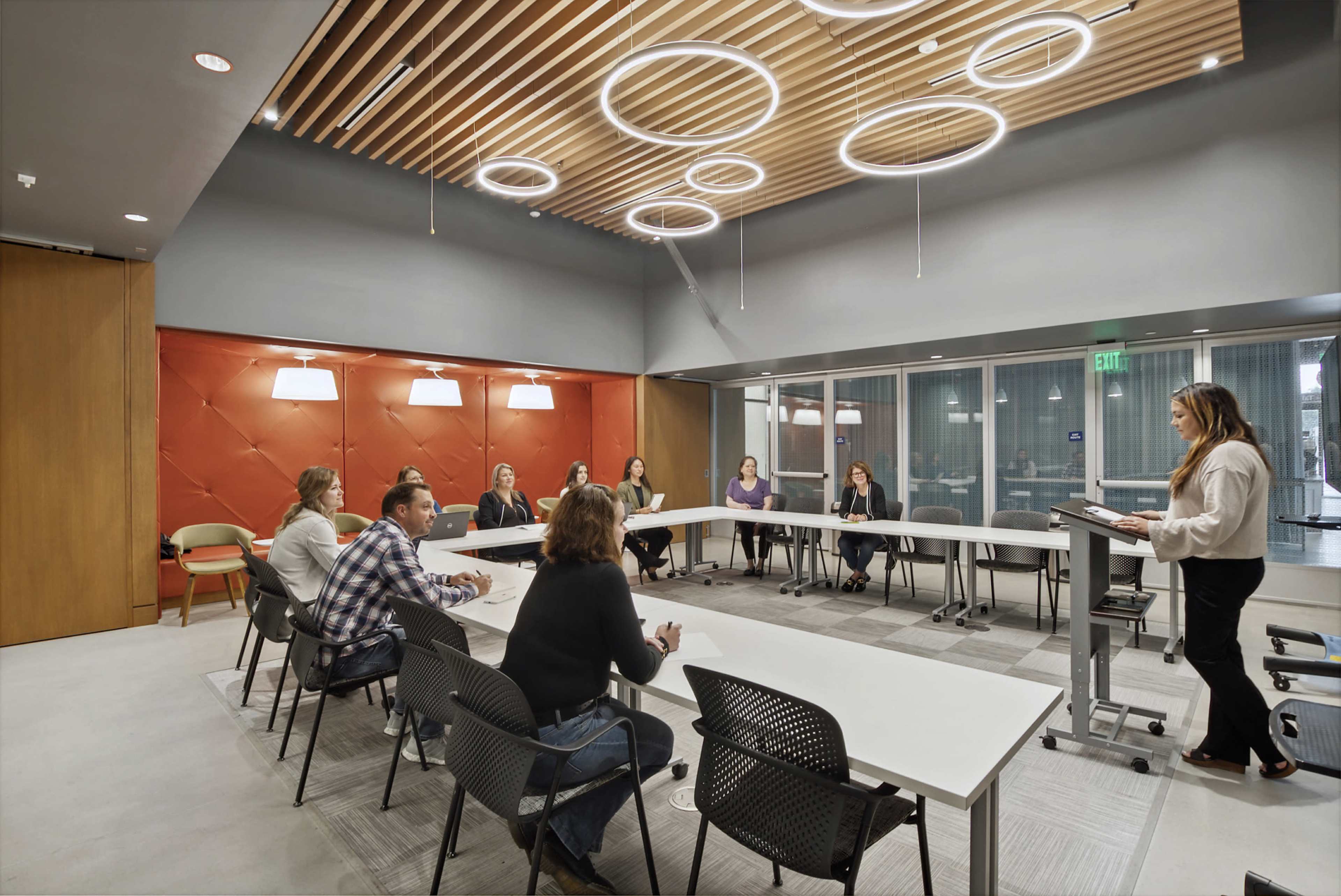 A group of people is seated around a long table in a modern conference room, while one person stands at the front presenting with a podium.