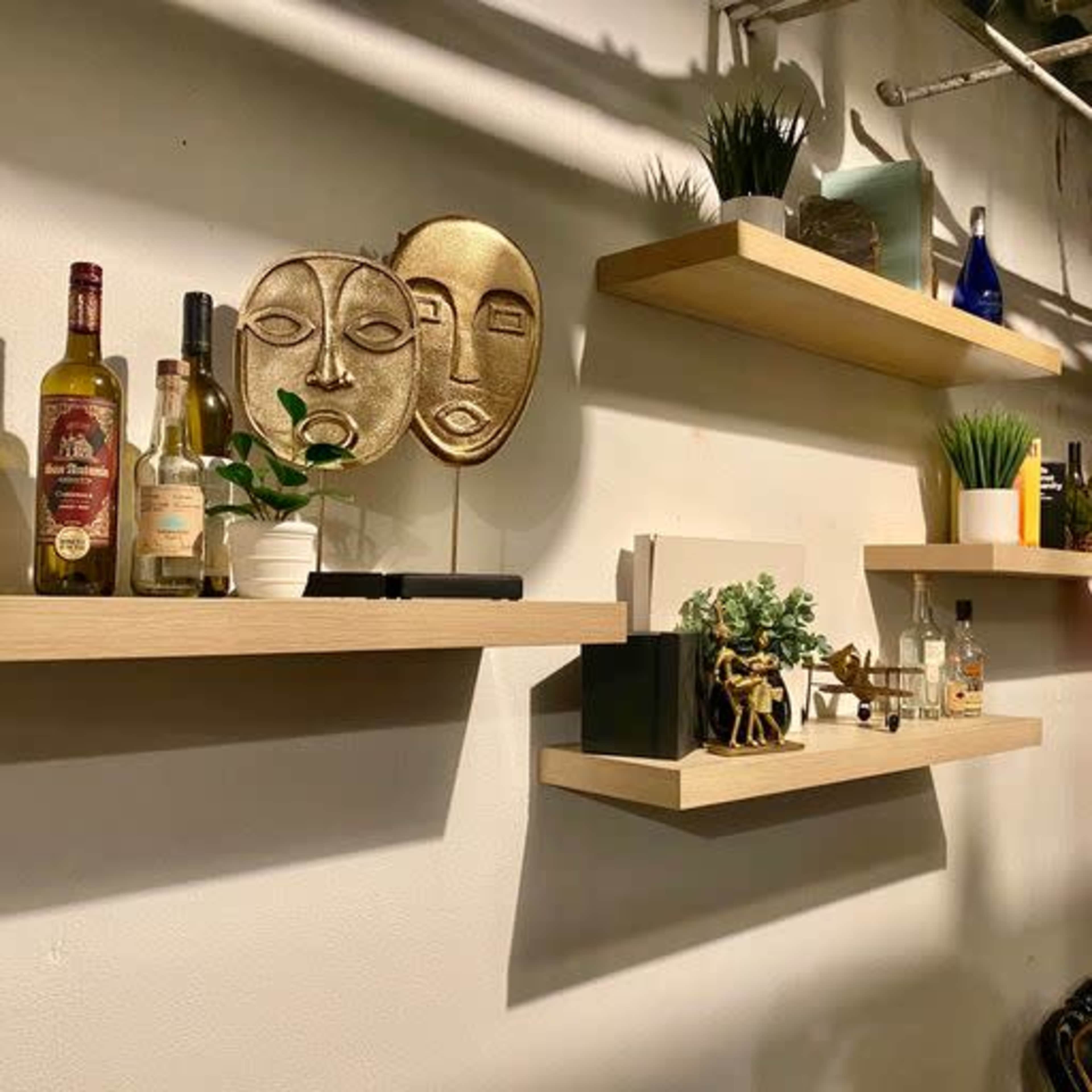 A wall with wooden shelves displaying bottles, plants, and decorative items, including two stylized face sculptures.