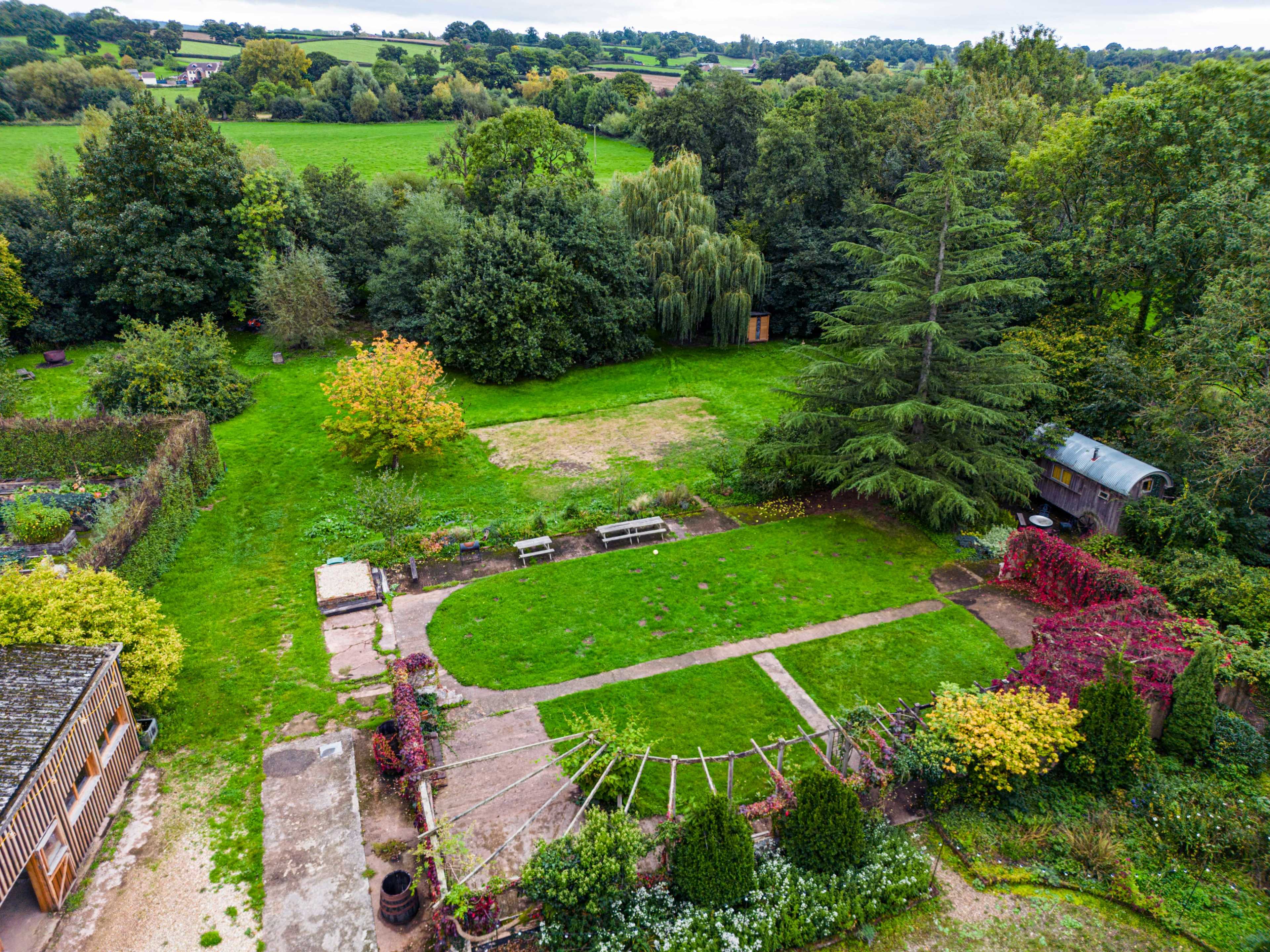 A landscaped garden with a paved pathway, benches, and a variety of trees and shrubs, surrounded by open green fields.