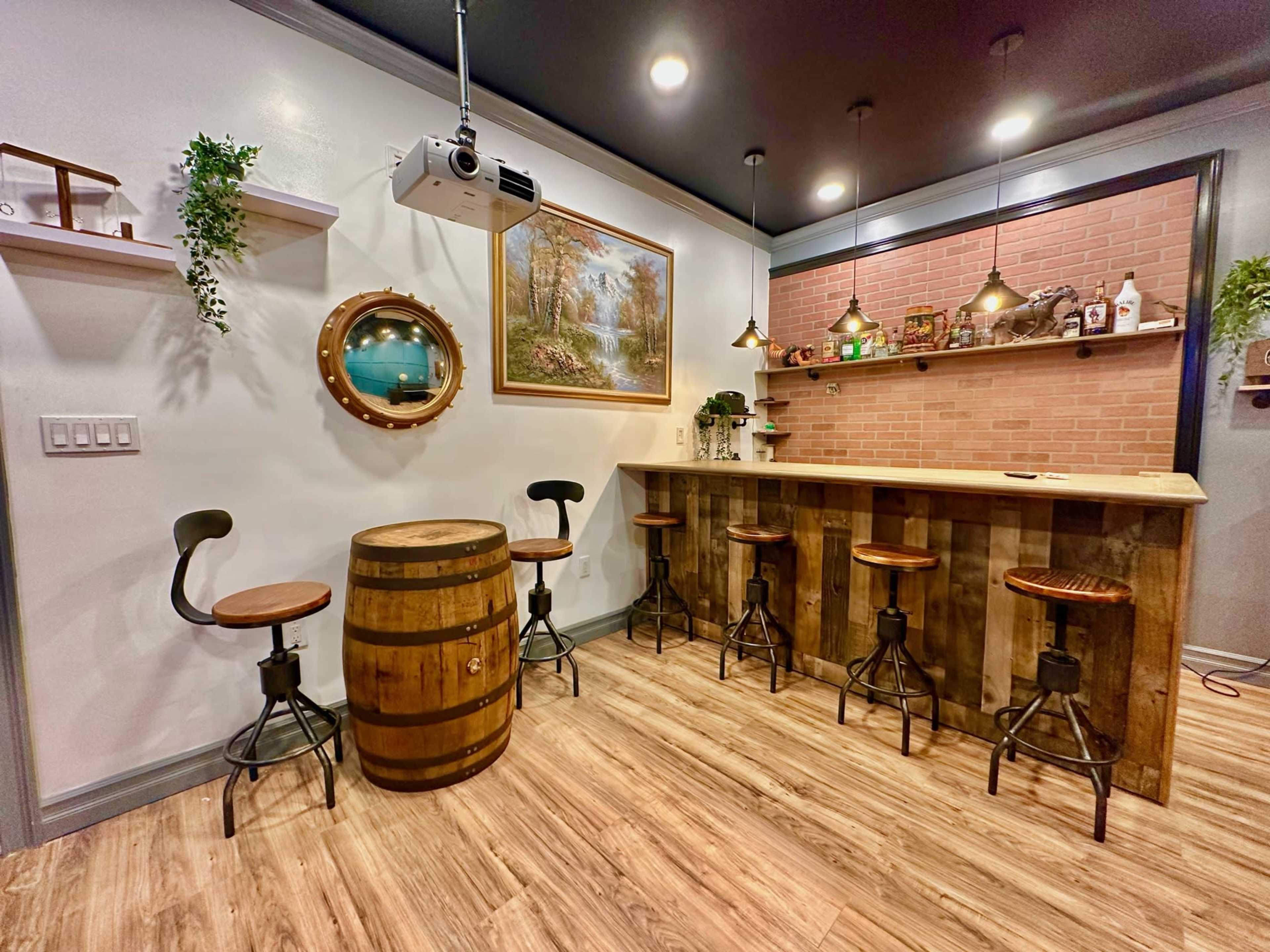 The image shows a cozy bar area with wooden stools surrounding a barrel table, a brick wall with shelves, and a projector mounted on the ceiling.