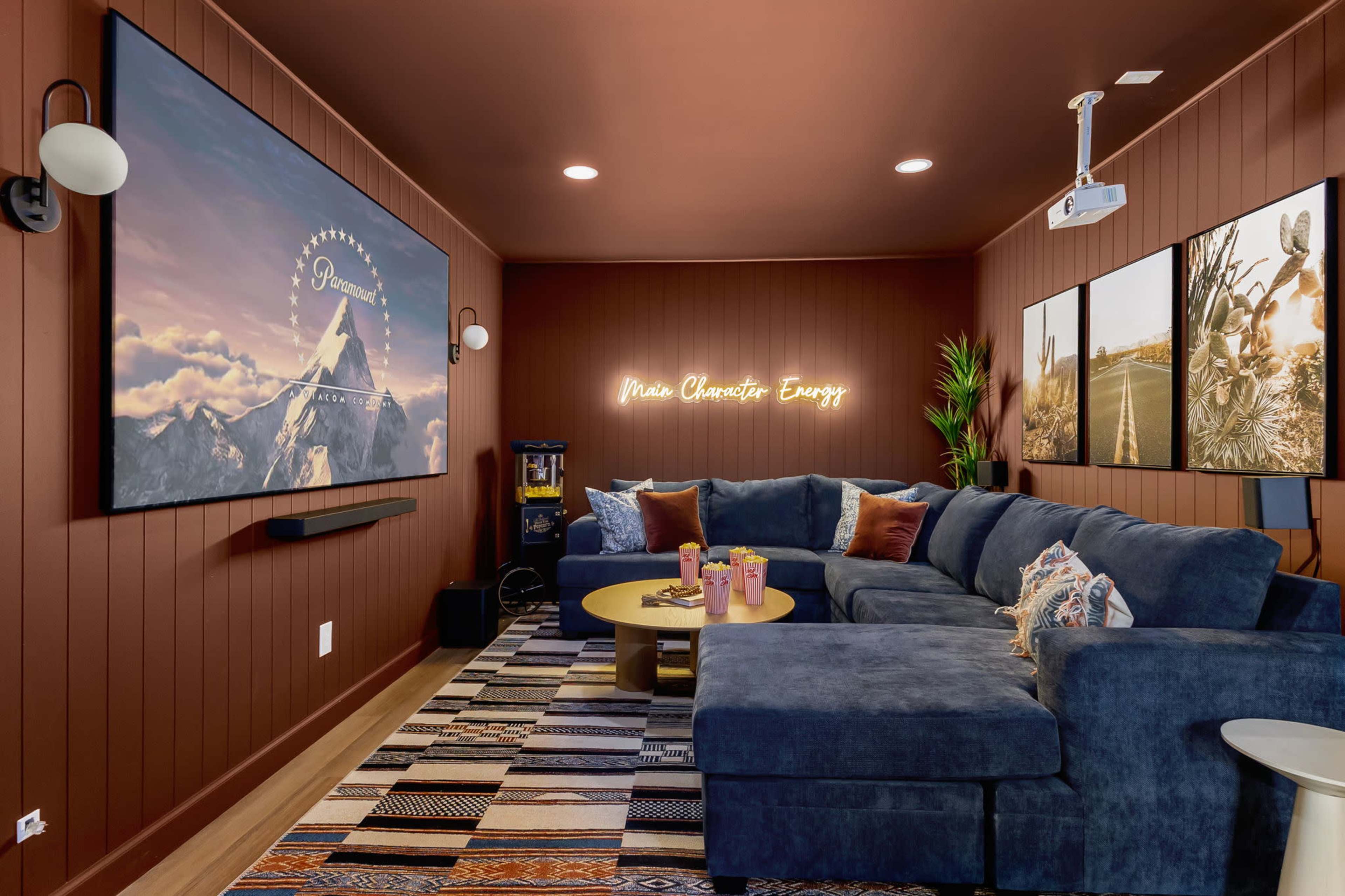 The image shows a cozy entertainment room featuring a large sectional sofa, a projector screen, and framed art on the walls.