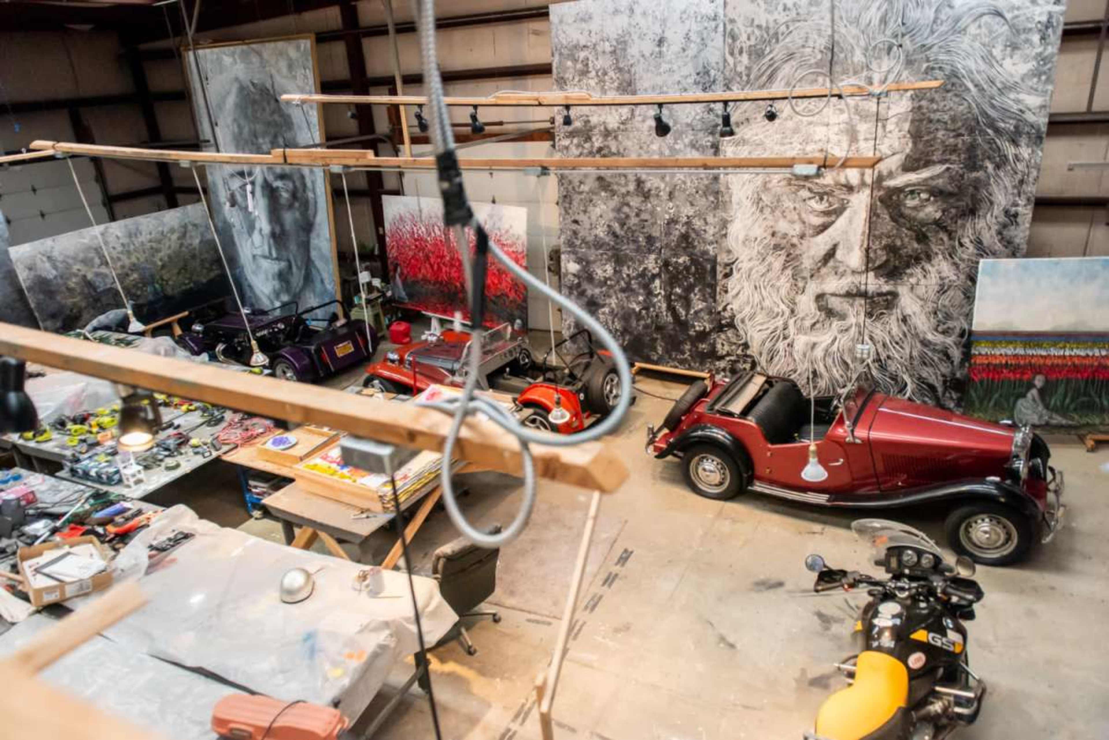 An artist's studio filled with large paintings, vintage cars, and various art supplies.