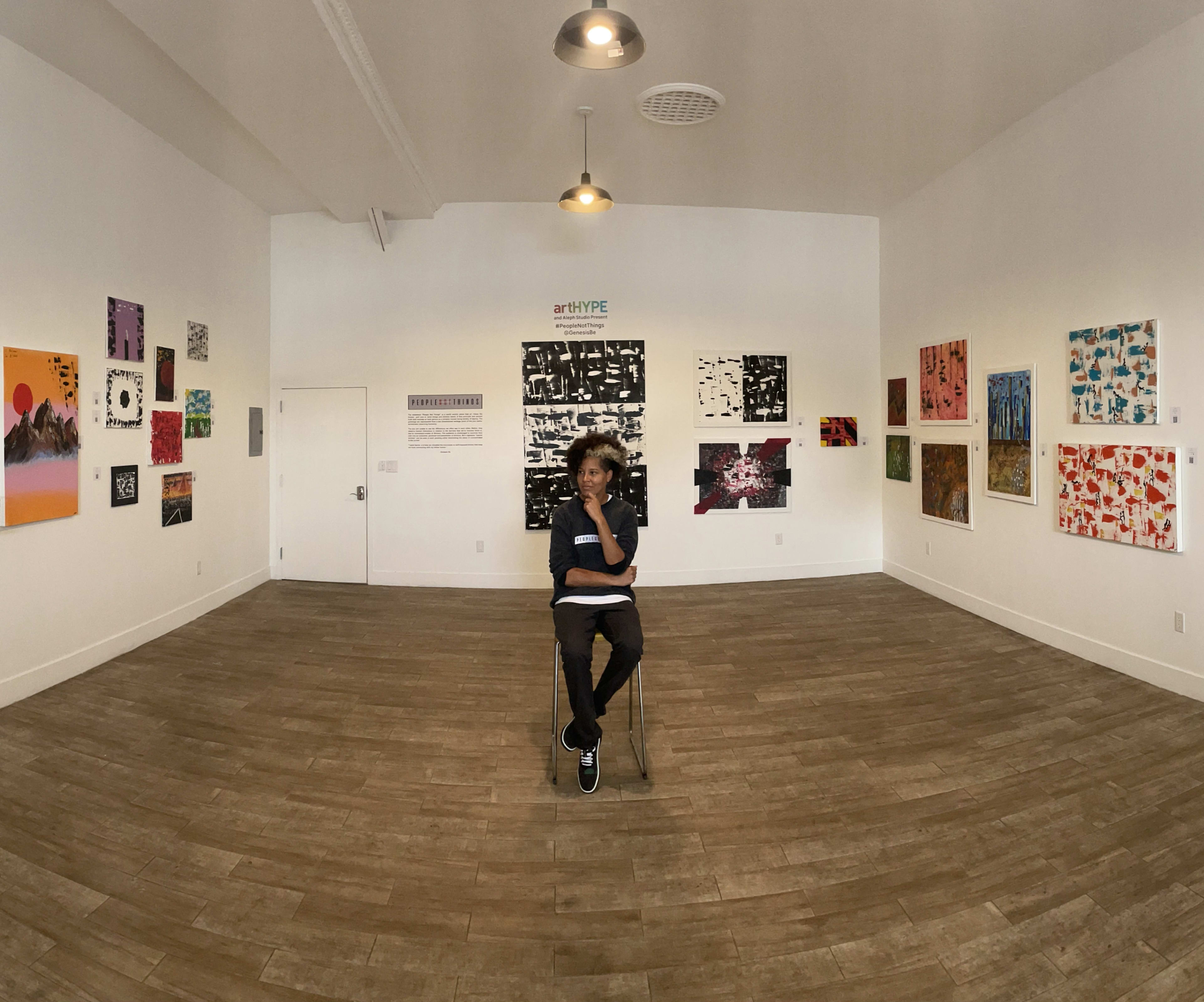 A person sits on a chair in the center of an art gallery surrounded by various artworks on the walls.