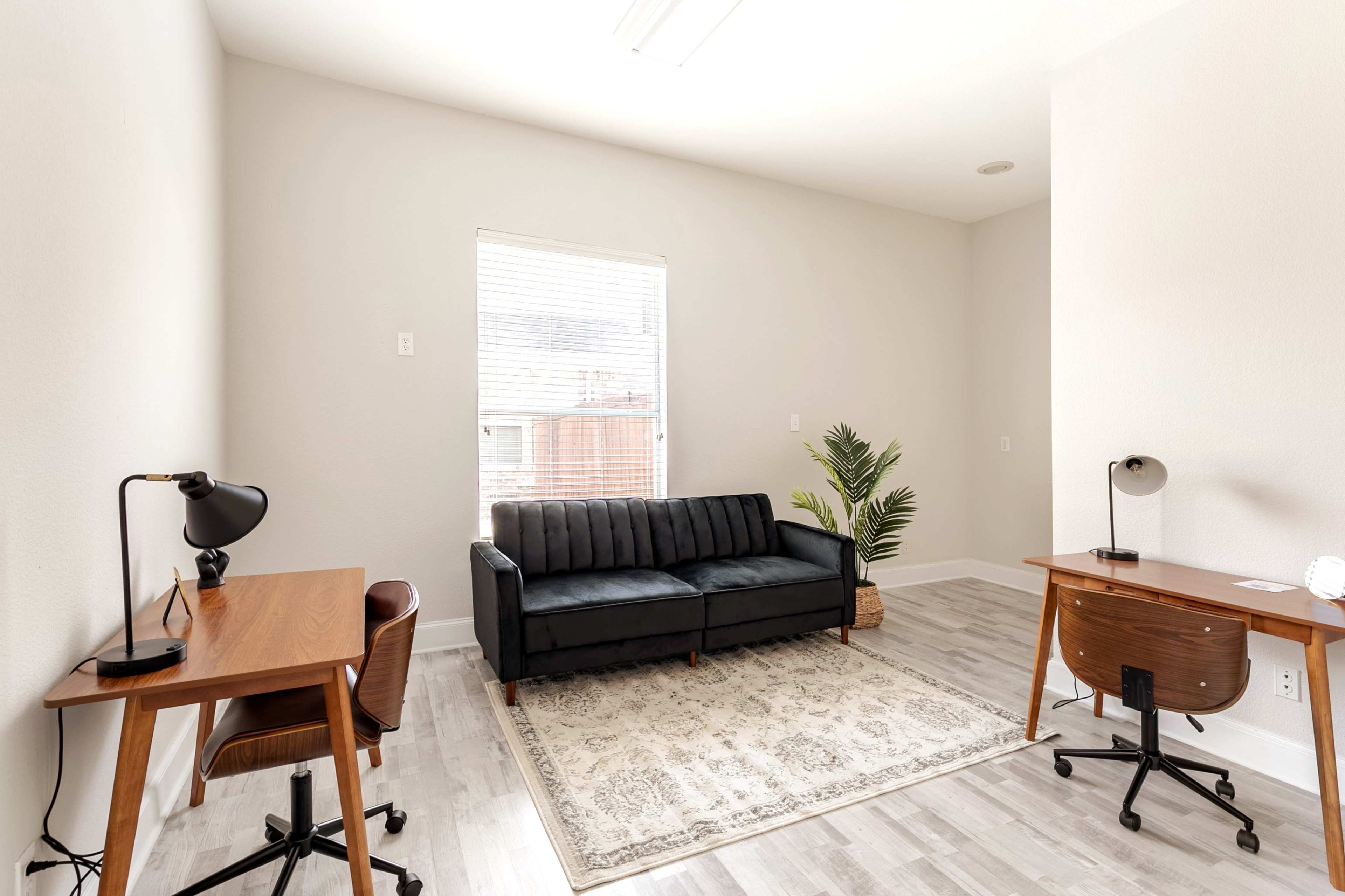 A light-colored room with two desks, a black sofa, a small plant, and a window.