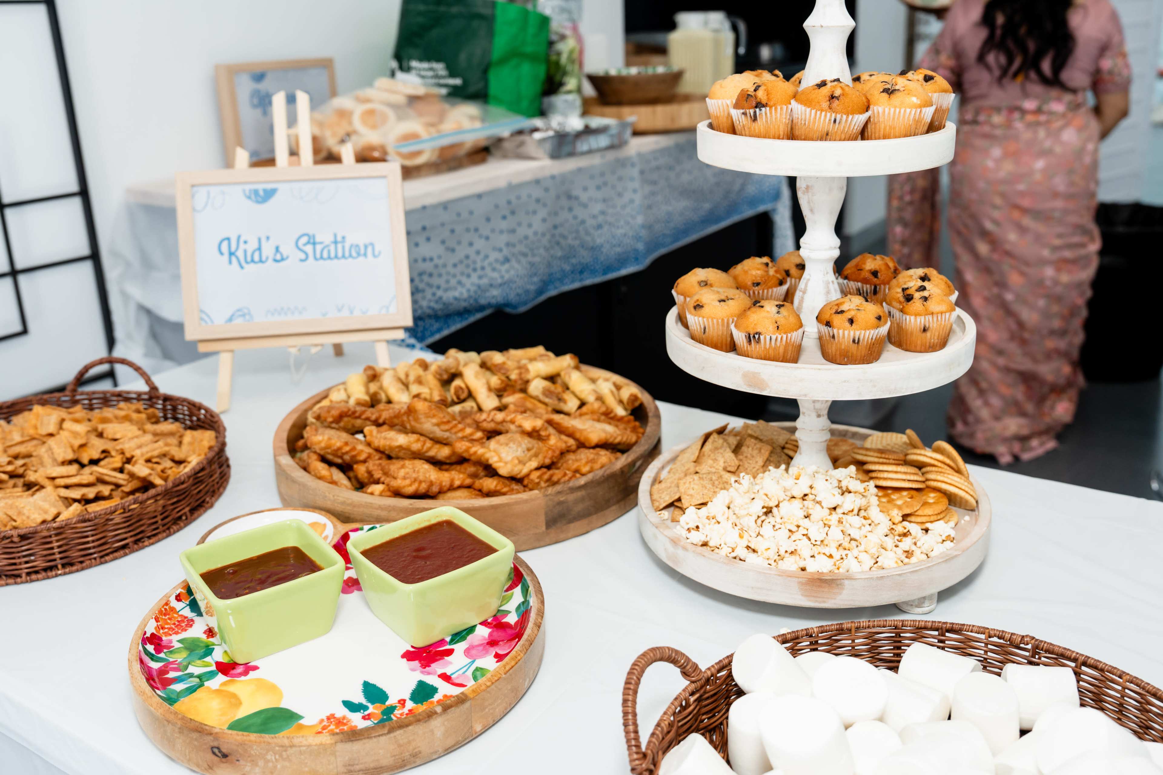 The image shows a variety of snacks served at a kid's station, including cookies, popcorn, crackers, and two bowls of dipping sauce.