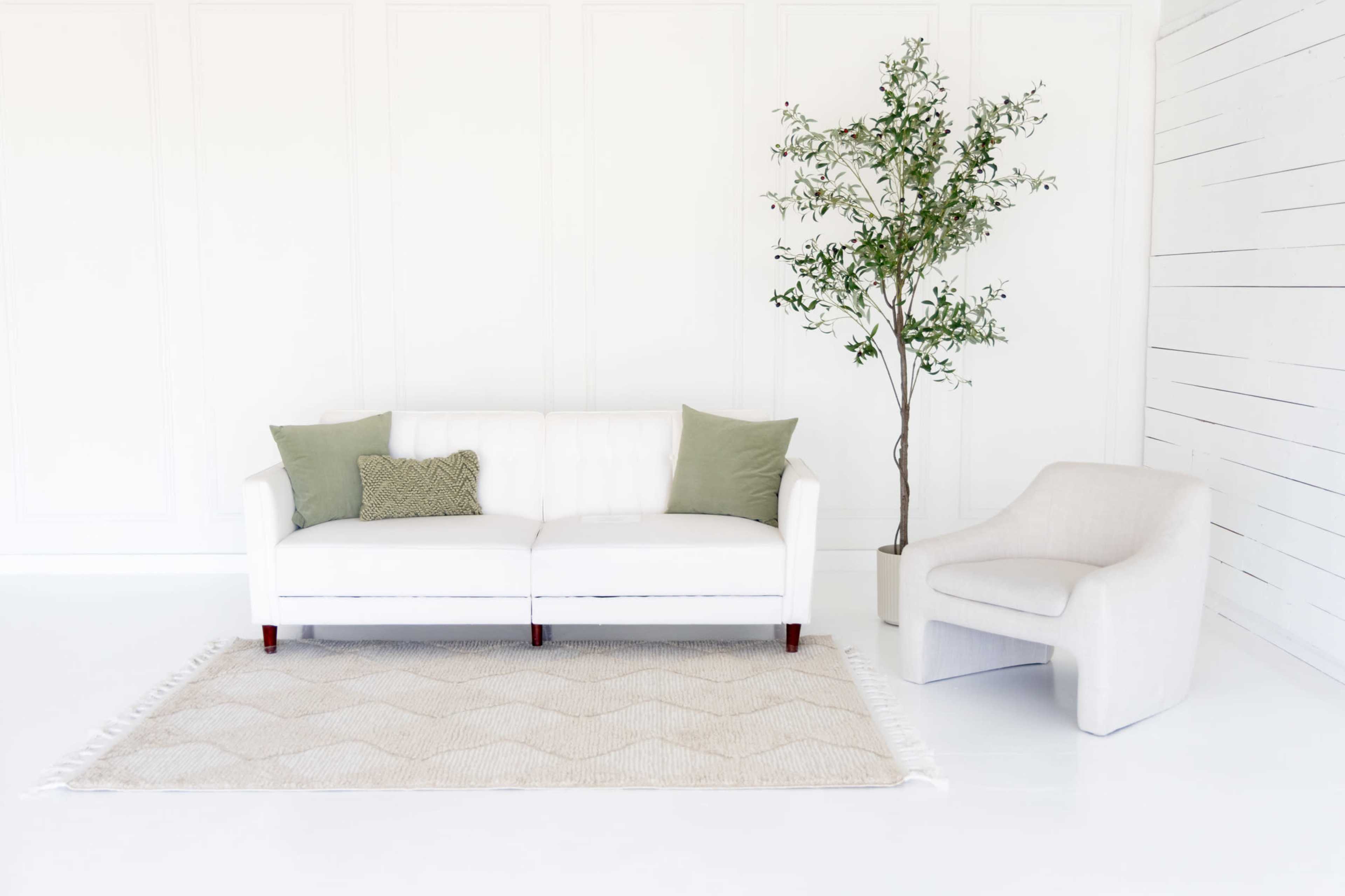 The image shows a white sofa with green cushions and a matching armchair, placed on a textured rug in a bright, minimalist room with a green plant beside the sofa.