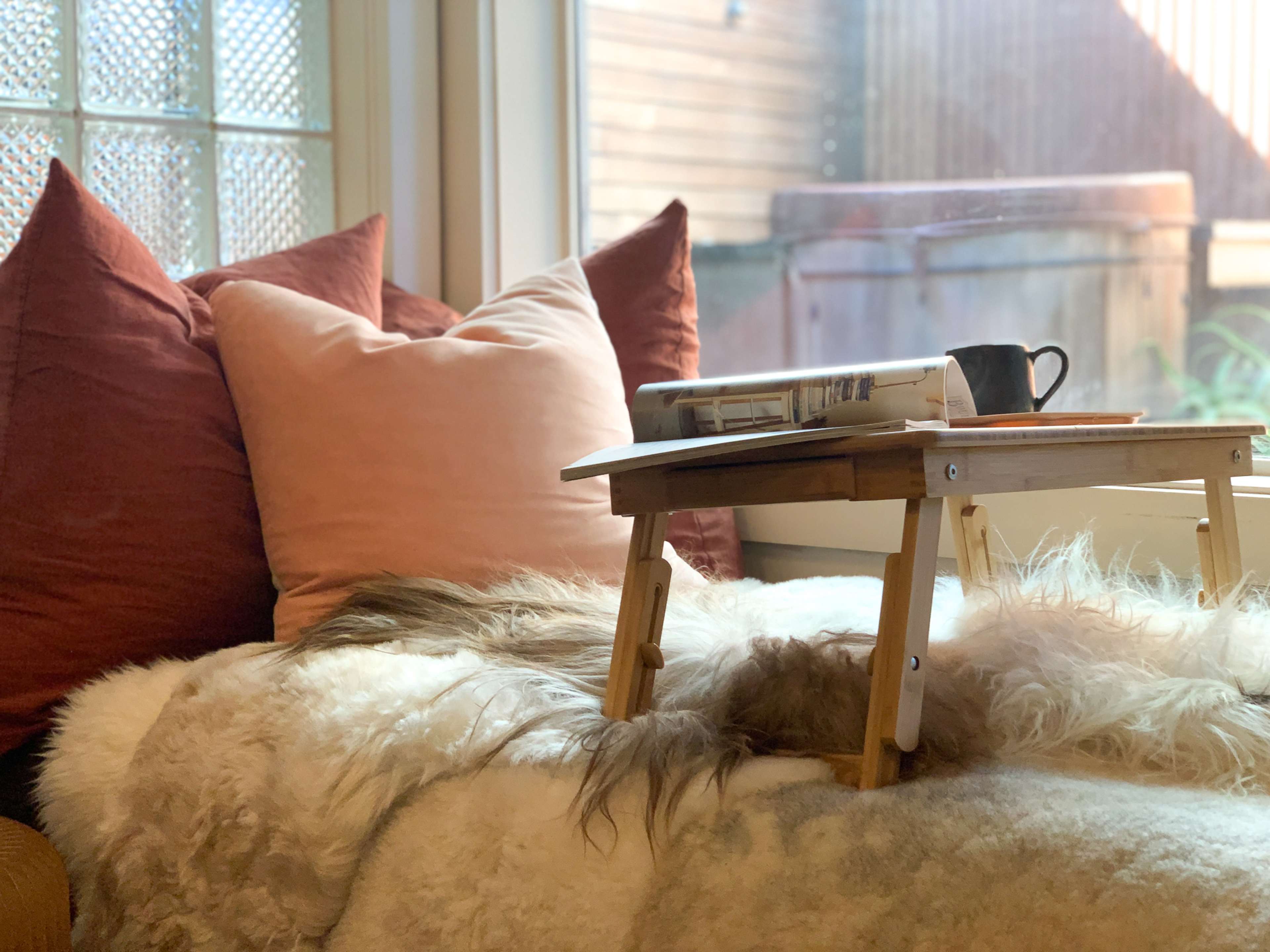 A small wooden table holds a newspaper and a mug in front of a cozy seating area with plush pillows and a soft throw blanket.