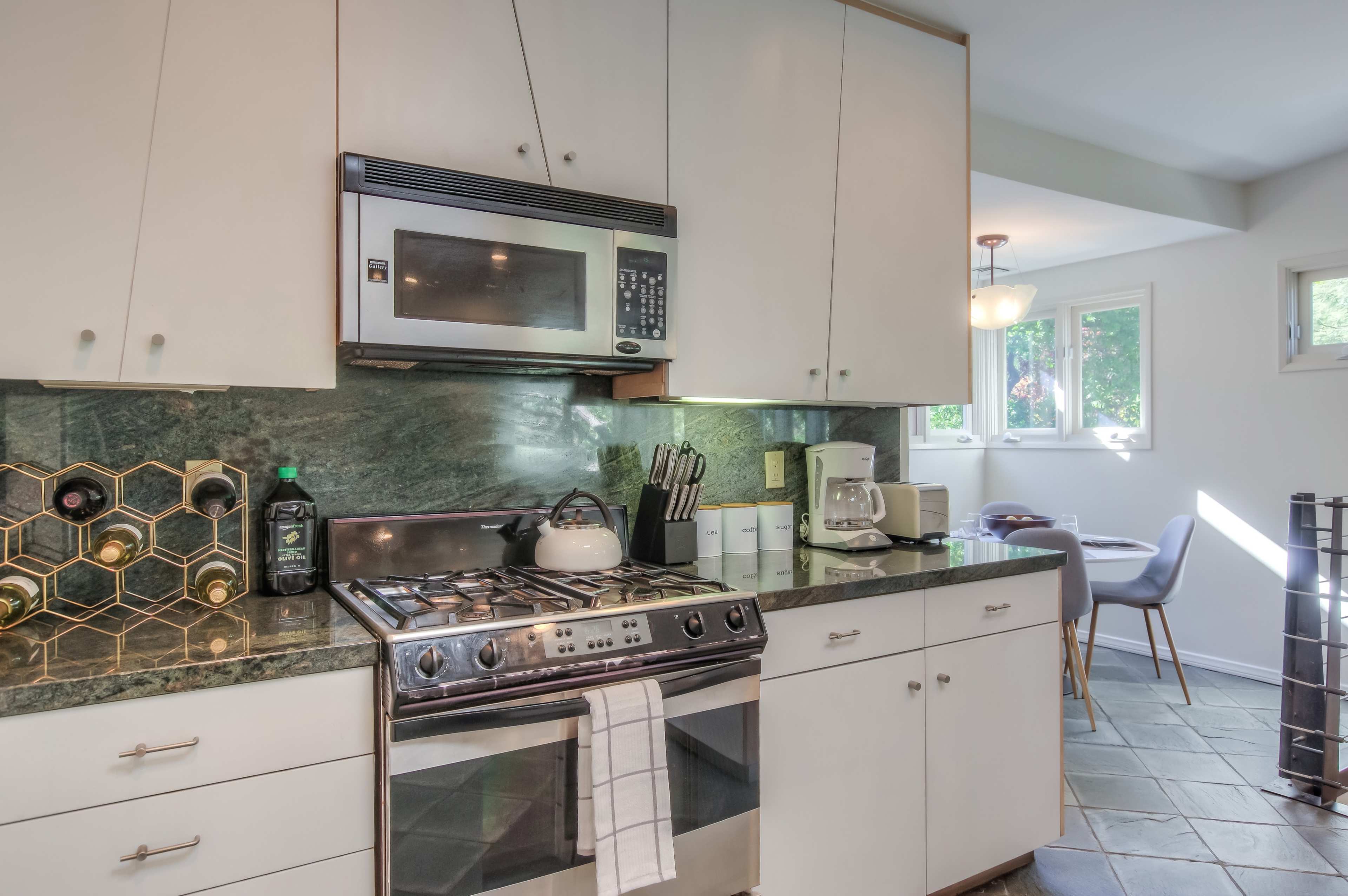 The image shows a modern kitchen featuring white cabinetry, stainless steel appliances, and a dark green stone countertop, with a dining area visible through a window.