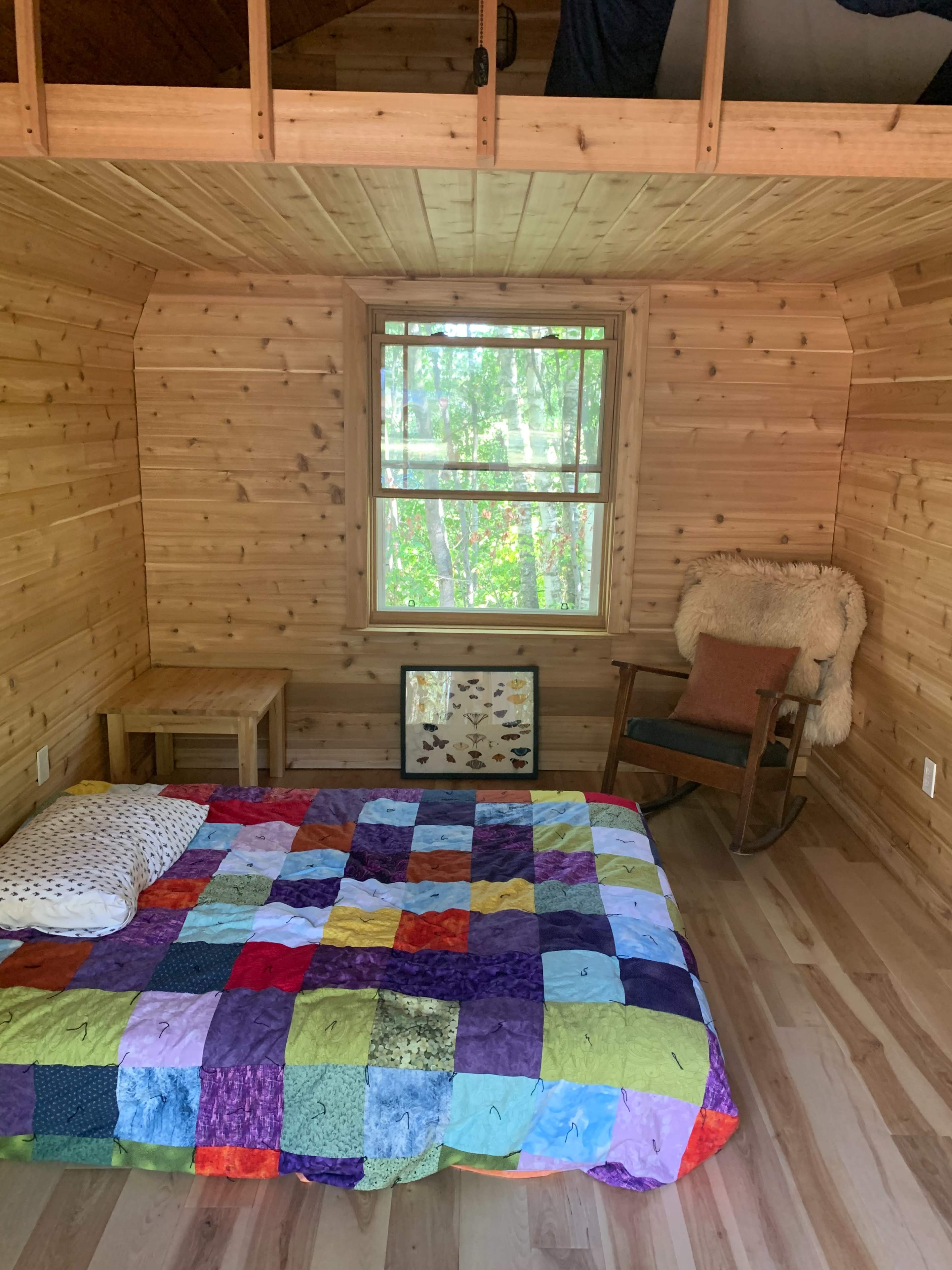 A wooden interior of a room with a quilt-covered bed, a small table, and a rocking chair near a window overlooking trees.