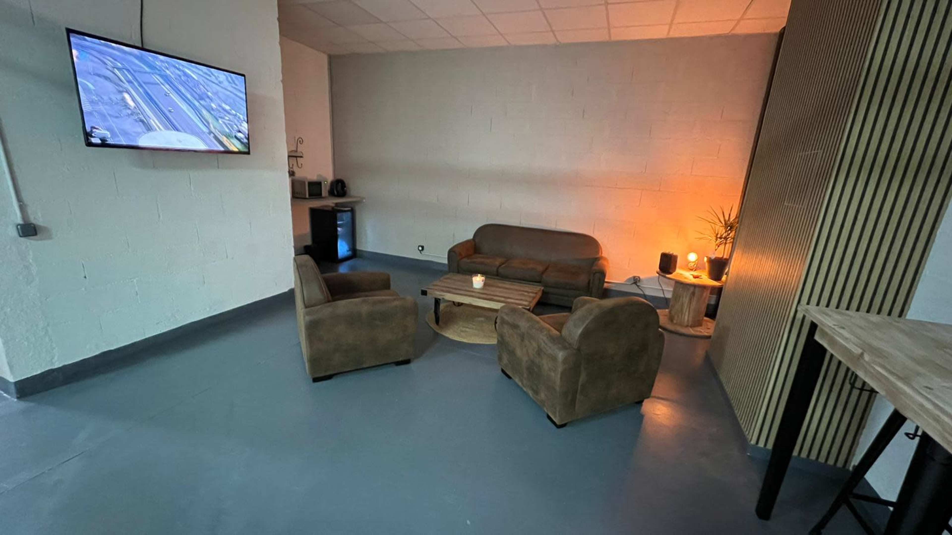A minimalistic lounge area with two brown couches, a wooden coffee table, and a small plant, illuminated by a warm light, and a television mounted on the wall.